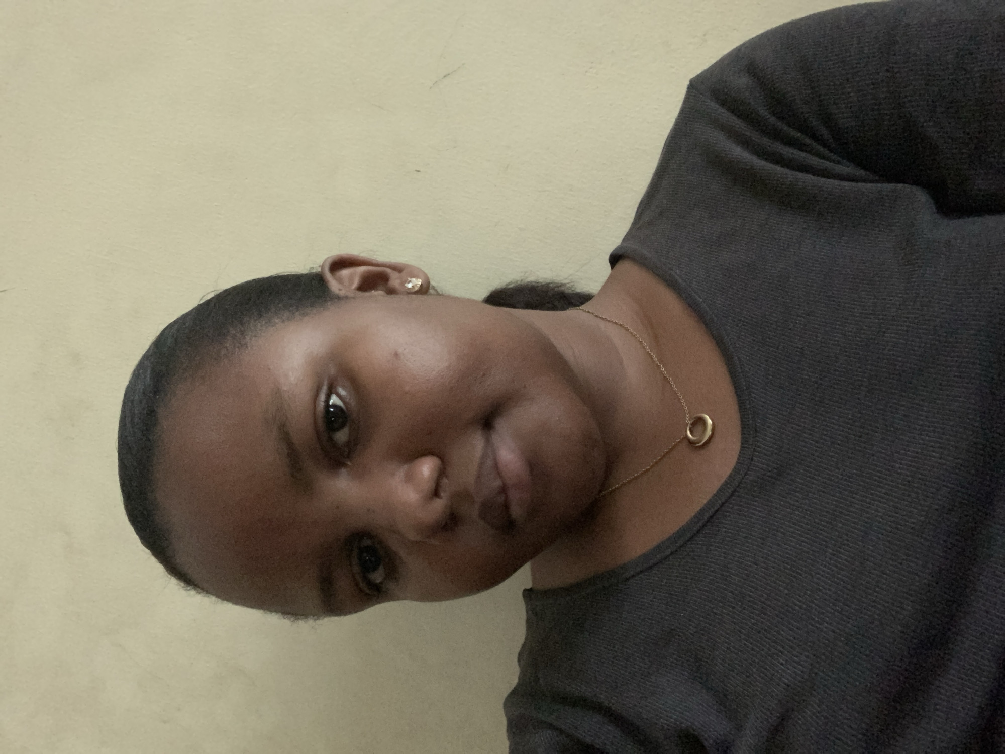 The image is a close-up portrait of a young adult woman with dark skin, positioned with a significant counter-clockwise tilt. She looks directly towards the camera with a neutral to slightly soft expression. Her dark hair appears pulled back from her face.
She is wearing a dark grey, long-sleeved top made of a finely textured fabric. Around her neck is a delicate gold chain necklace with a small, simple gold circular pendant. In her visible left ear (viewer's right), she wears a clear or light-colored stud earring.
The background consists of a plain, light yellow or cream-colored wall, which shows some minor smudges or discolorations in various areas. The lighting is soft and even, suggesting an indoor setting. No specific activity beyond posing for the portrait is depicted. There is no visible text in the image. The provided context indicates the location is Dar es-Salaam, Tanzania, though this is not discernible from visual cues within the image itself.