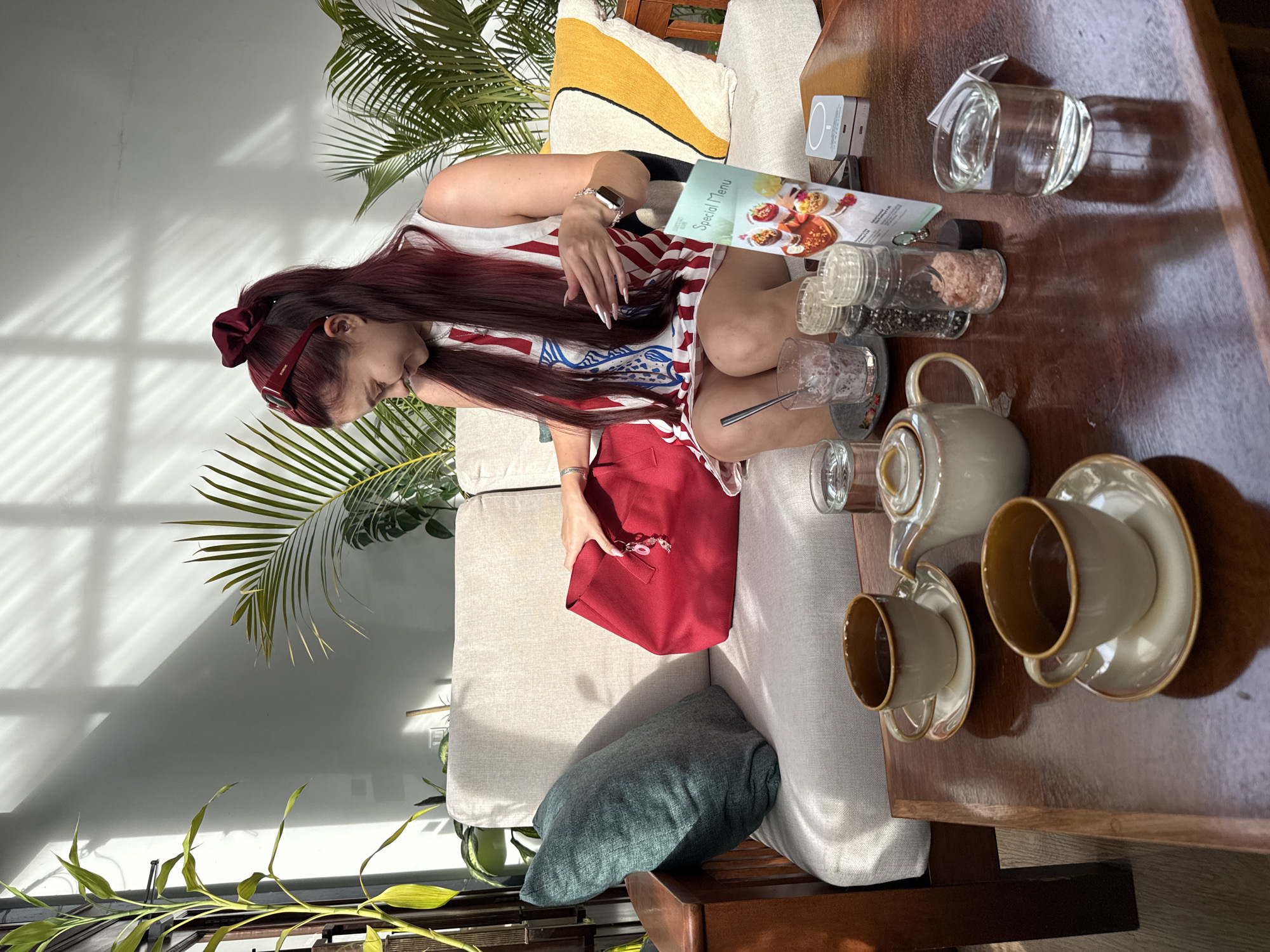 A woman with long, dark red hair pulled into a high ponytail and adorned with a red scrunchie and sunglasses, sits on a sofa in an indoor cafe, possibly in Khan Daun Penh, Cambodia. She is wearing a white sleeveless dress with red and blue vertical stripes. She is looking down and to her right, with her left hand reaching into a large red tote bag. On the wooden table in front of her are two teacups and saucers filled with dark liquid, a teapot, two salt and pepper shakers, a glass of water, a glass with a spoon in it, and a menu. The cafe has large windows that cast striped shadows on the wall, and several potted plants are visible, including a tall bamboo plant on the left and palm-like plants in the background. The sofa is light-colored with a teal pillow and a yellow and white striped pillow.