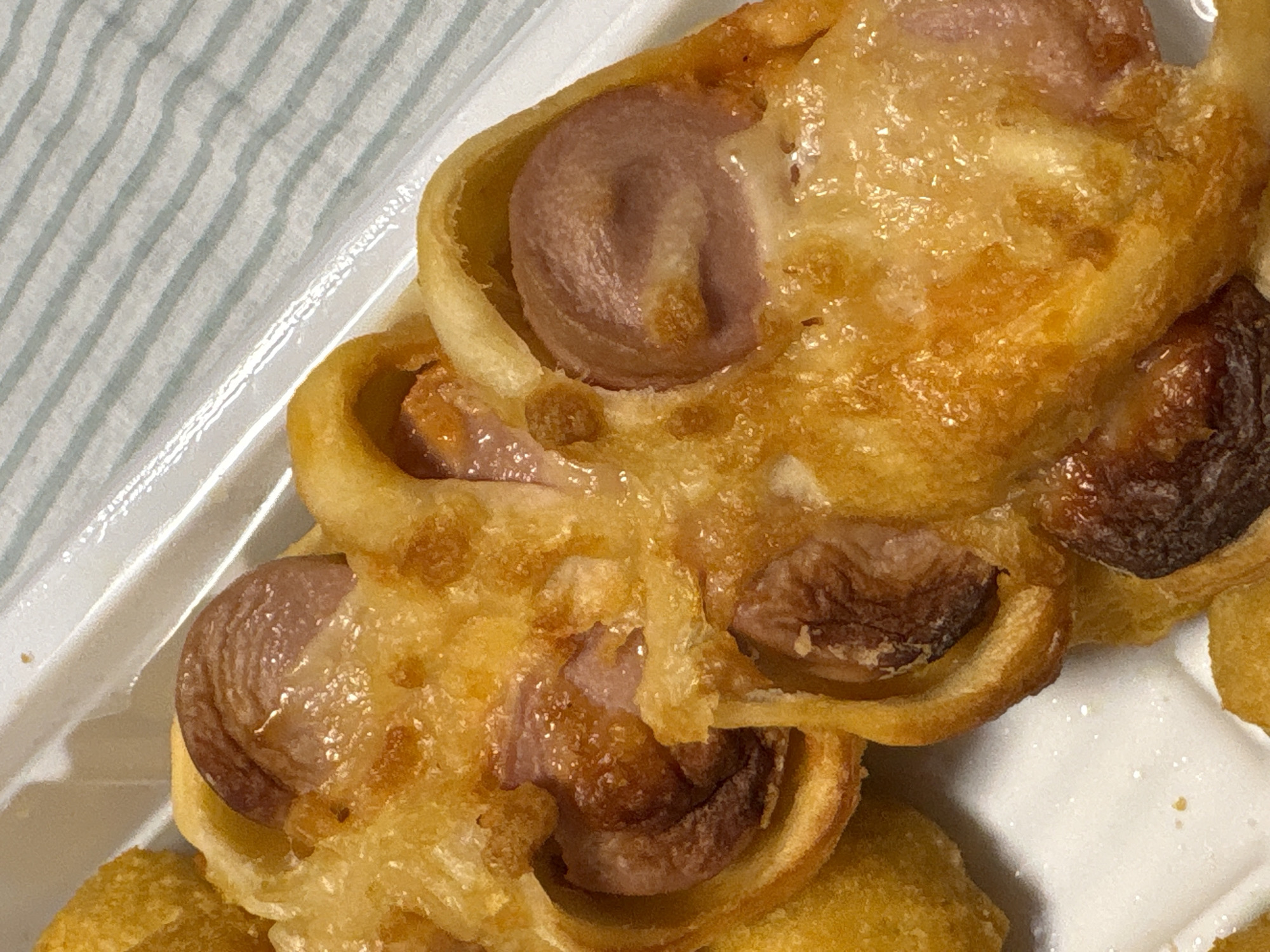 A close-up shot reveals a sausage roll pastry on a white plastic container. The pastry encases sliced sausages, and the entire roll has a glazed, golden-brown appearance, indicating it has been baked or fried. The setting appears to be indoors. No individuals are visible in the image. The container sits on a surface with pale, horizontal stripes. The specific location within Прикамский, Russia is indeterminable from the image.