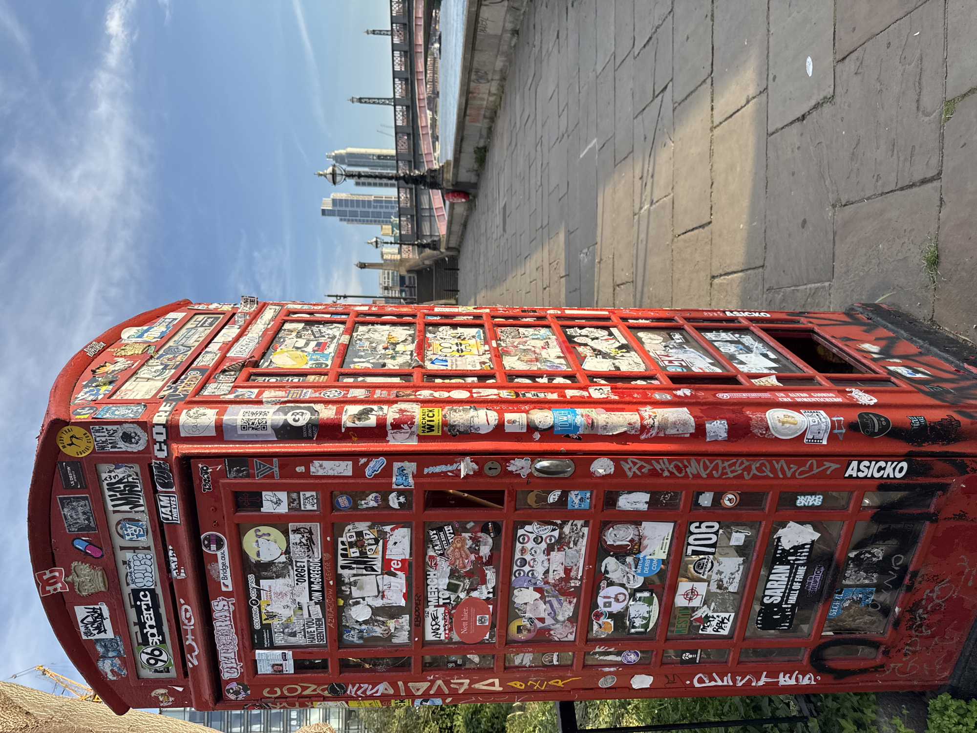 A red, graffiti-covered telephone booth stands on a paved walkway beside the River Thames in London. The booth is covered in stickers and graffiti. In the background, a bridge spans the river, with modern buildings visible on the far bank. The sky is blue with scattered clouds. The walkway is made of large stone slabs. There are no people in the image.