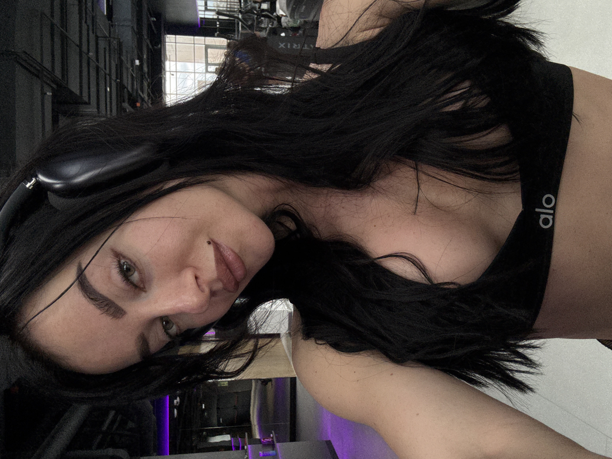 A woman is in a gym. She is wearing a black sports bra with "alo" on the strap and headphones. Her long black hair is partially obscuring her face. The gym has weight machines visible in the background. The location is potentially in the city of Krasnoyarsk, Russia.