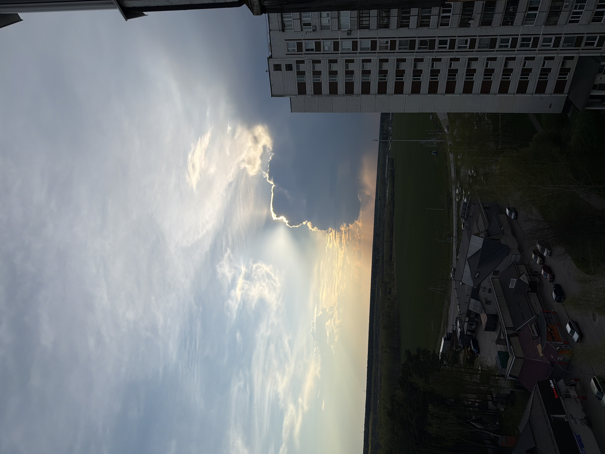 The image captures a view from a high vantage point, likely a building in Chernogolovka, Russia. The composition is divided between sky and land. In the sky, there are clouds with sun rays breaking through. There is also a part of the top of a building, implying the photographer may be on top of it. The land shows a grassy area, buildings, and possibly a road with some vehicles, though the details are indistinct due to the distance.