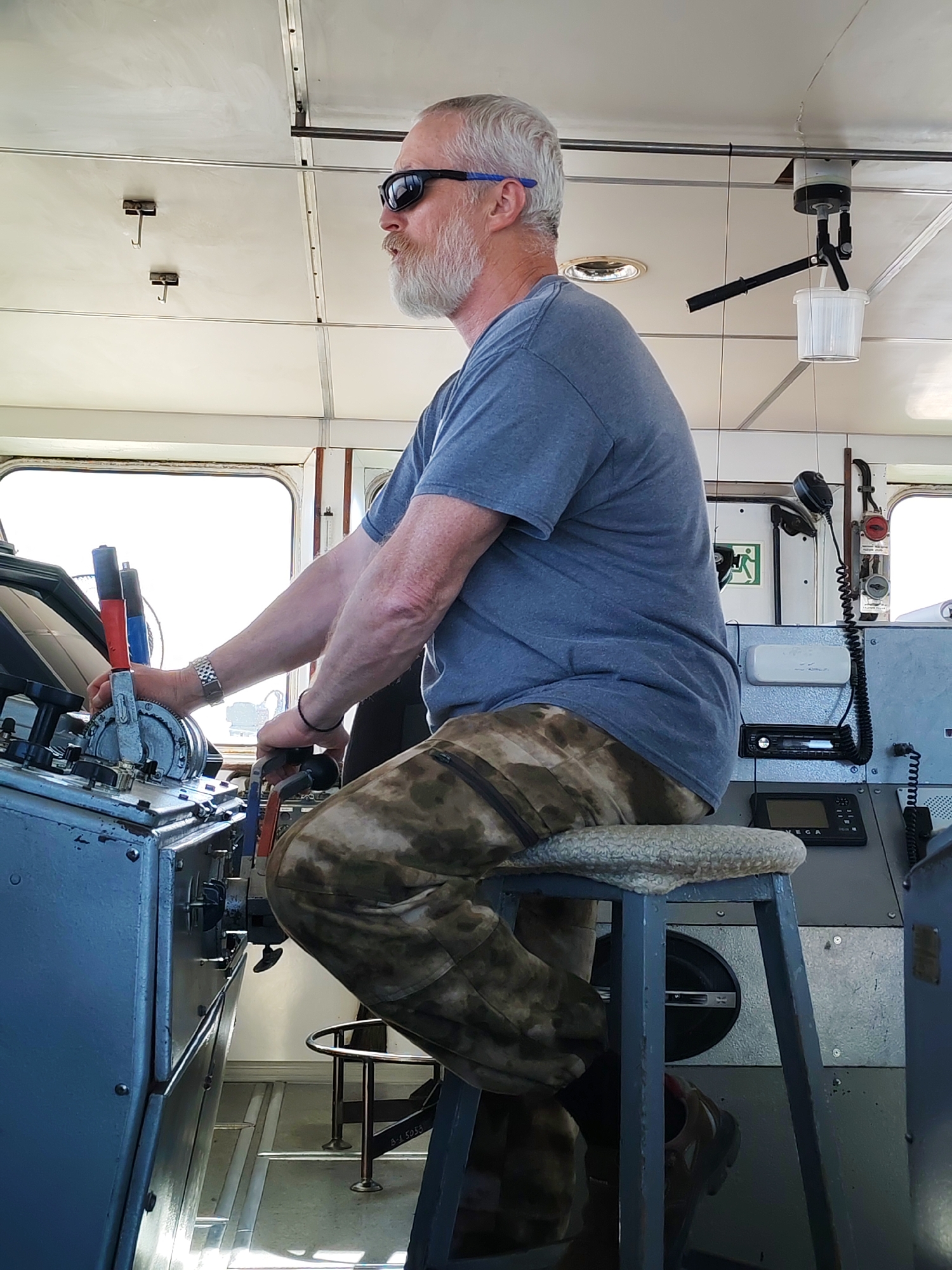 An elderly male with grey hair and a grey beard, wearing dark sunglasses with blue accents, a grey t-shirt, and camouflage-patterned trousers, is seated and operating controls in what appears to be a ship's bridge, located in the city of Unknown, Unknown. He is positioned in profile, facing the right side of the frame. His left hand manipulates a metal lever with a red top on a grey control panel to his left, while his right hand operates a black control knob or handle. A silver-toned watch is visible on his left wrist and a dark bracelet on his right wrist.
The subject is seated on a stool with a light-colored, woven cushion and a dark metal frame. To his left, the control panel is light grey and features multiple levers (one also visible with a blue top), dials, and other controls. To his right, another grey console holds communication equipment, including a black coiled cord and microphone, along with various electronic displays, one showing the brand "OMEGA".
The interior features a light-colored, paneled ceiling with metal rails, several metal hooks, and a circular overhead light fixture. A white cylindrical apparatus with black attachments and a clear plastic bucket suspended from it hangs from the ceiling on the right. Large windows on the left and right sides of the space allow bright ambient light, rendering the exterior view overexposed. A green "exit" sign is visible on a white surface in the mid-ground. The floor is light-colored, featuring horizontal metal strips, and reflects the ambient light.