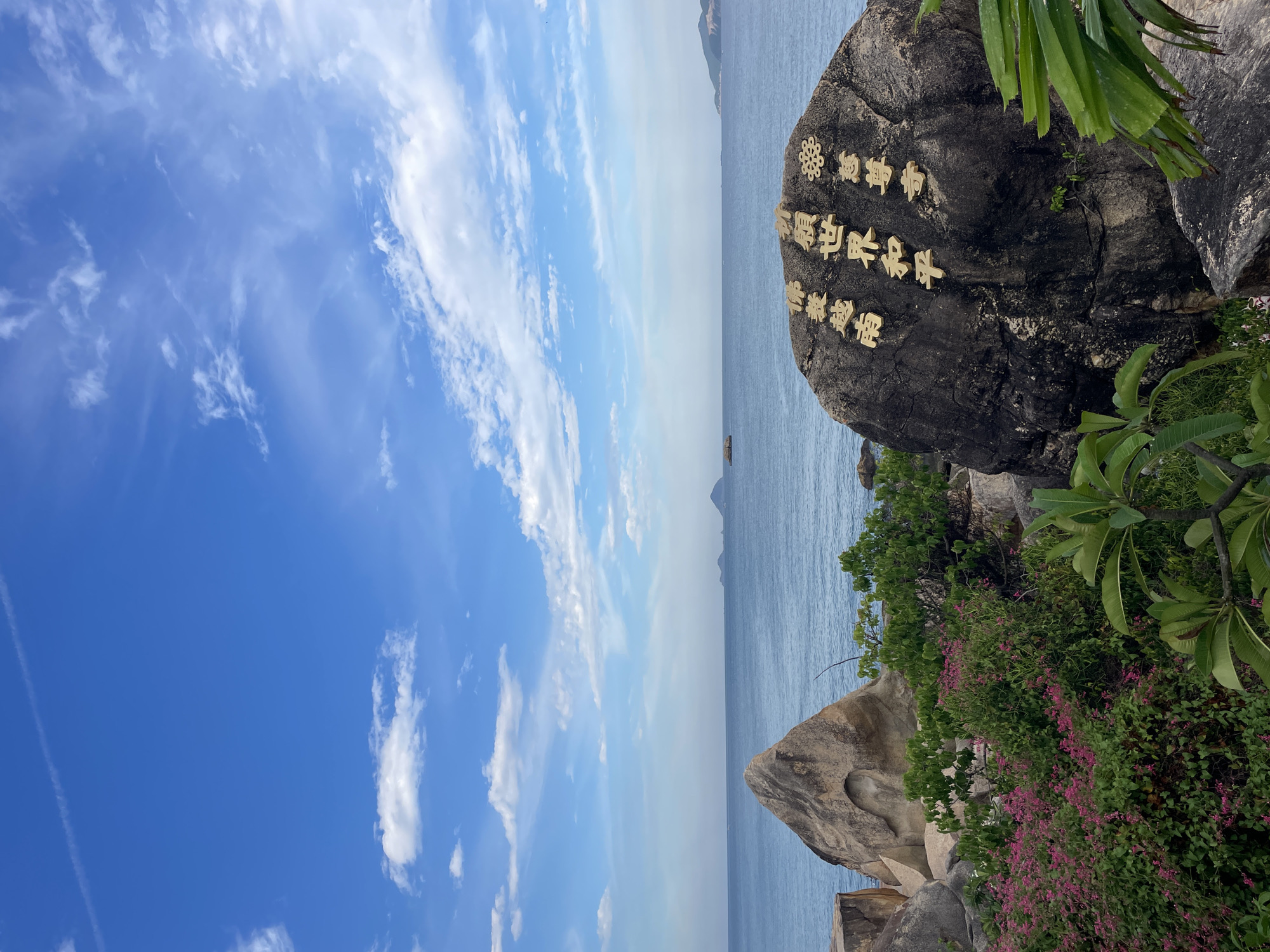 The image depicts a coastal scene in Nha Trang, Vietnam, featuring a large, dark rock inscribed with gold lettering. The rock is positioned in the foreground, dominating the right side of the frame. The golden characters appear to be in an Asian script, likely Chinese or Vietnamese. Behind the rock, the calm, blue sea stretches to the horizon, with several islands visible in the distance. The sky is a vibrant blue, dotted with scattered white clouds. To the left of the large rock, a lighter-colored, triangular rock formation is visible, and in front of these rocks, lush green foliage and vibrant pink flowers create a naturalistic setting. The overall atmosphere is serene and picturesque, indicative of a tourist or scenic location.