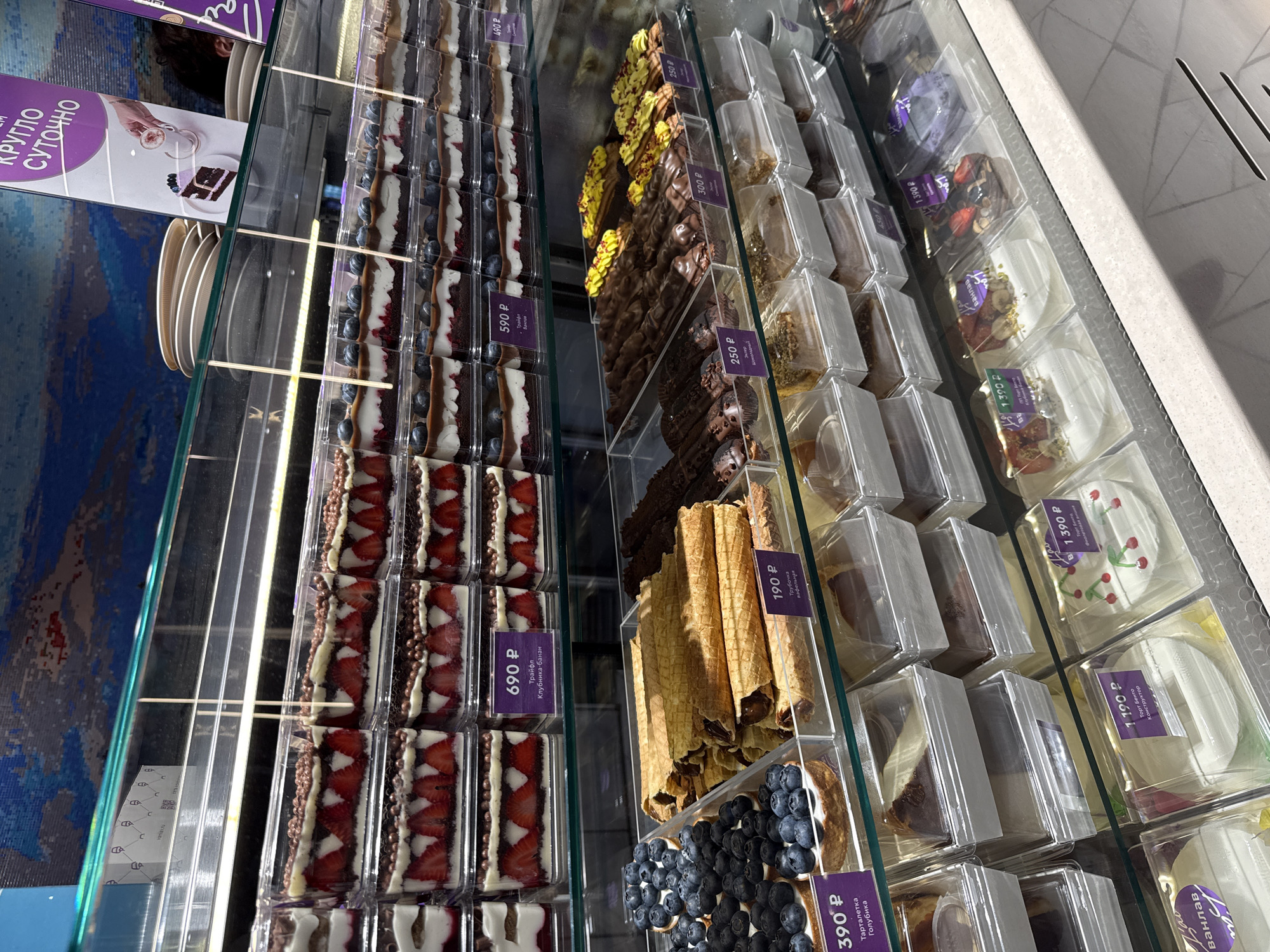 Here's a detailed description of the image:
**Scene:** The image shows a display case filled with various desserts, likely in a bakery, cafe, or grocery store in Krasnoyarsk, Russia. The desserts are packaged in clear plastic containers and arranged neatly on shelves.
**Desserts:** The variety of desserts includes:
* Truffle-like desserts with strawberry, and blueberry toppings.
* Chocolate-covered desserts.
* Waffle tubes.
* Tarts with blueberries.
* Bento cakes.
* Other various cakes and pastries.
**Pricing and Labels:** Each dessert has a small purple label indicating the price in Russian rubles (P). The labels also include the name of the dessert, such as "Трайфл Клубника-банан" (Trifle Strawberry-banana), "Тарталетка Голубика" (Tartlet Blueberry), "Трубочка вафельная" (Waffle tube) and "Торт Бенто" (Bento Cake).
**Setting:** The display case has a glass front and interior lighting to showcase the desserts. The background is slightly blurred, but suggests a commercial environment.
**Additional elements:** On the left of the image, a sign with text "КРУГЛО СУТОЧНО" is visible.
The overall scene is a well-organized and appealing display of desserts, inviting customers to purchase them.