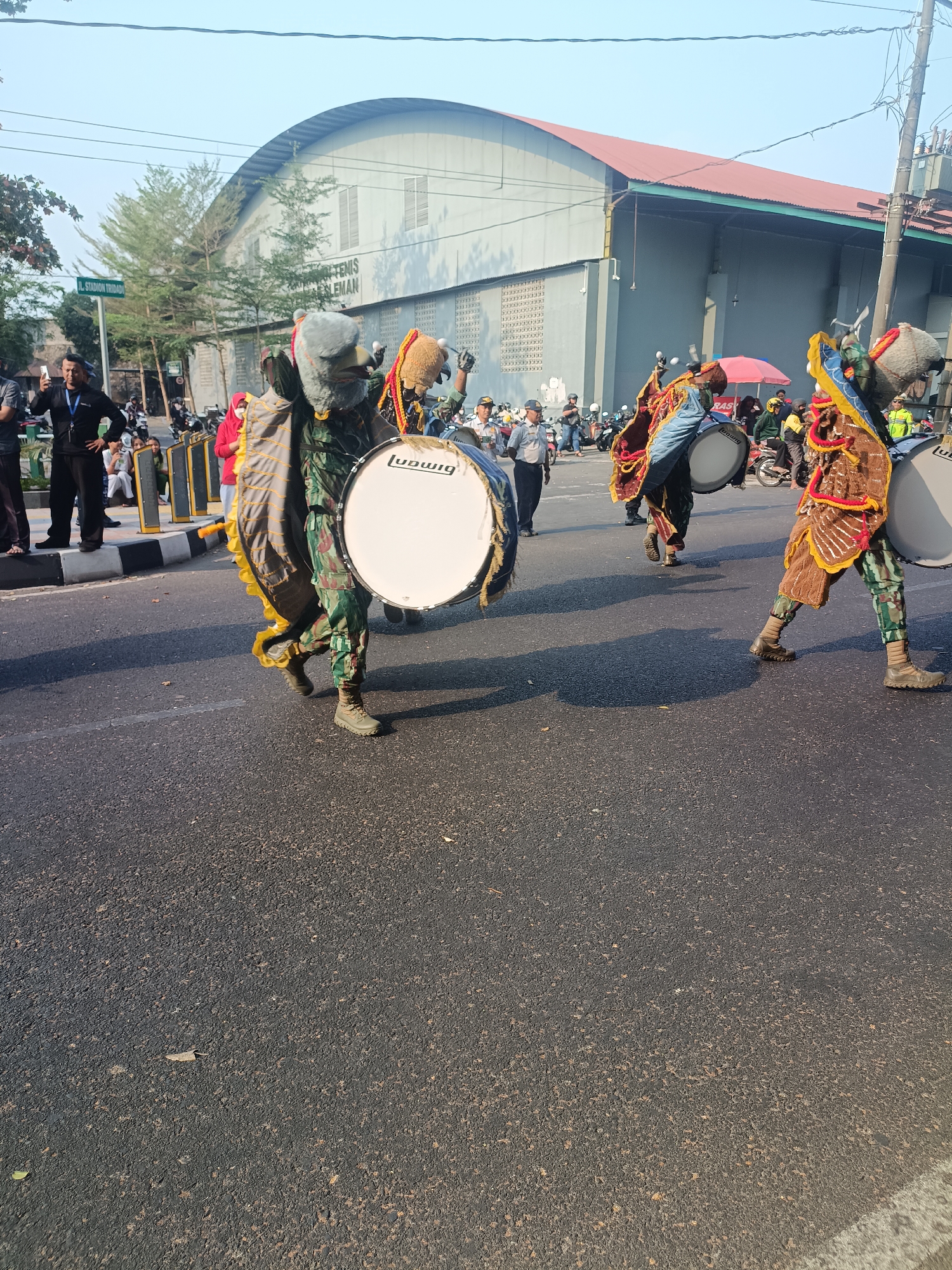 The image depicts a street scene in Sleman, Indonesia, likely during a parade or cultural event. In the foreground, three individuals dressed in elaborate, colorful costumes, complete with what appear to be animalistic headdresses, are marching and carrying large bass drums. The costumes feature camouflage-patterned trousers, embellished vests, and fringed adornments in shades of brown, yellow, and red. The drums are white with the "Ludwig" brand name visible on one.
Behind the drummers, a crowd of spectators lines the street. Many are on foot, while others are on motorcycles, suggesting a lively public gathering. A street sign with the text "JL. STADION TRIDARMA" is visible on the left, indicating the location is near a stadium. The background features a large, light-grey building with a red roof, possibly a sports arena or convention center, with the words "TENIS" and "SLEMAN" partially visible on its facade. The sky is clear and blue, and the sunlight casts shadows on the asphalt road. The environment appears to be an urban street setting on a bright, sunny day.
