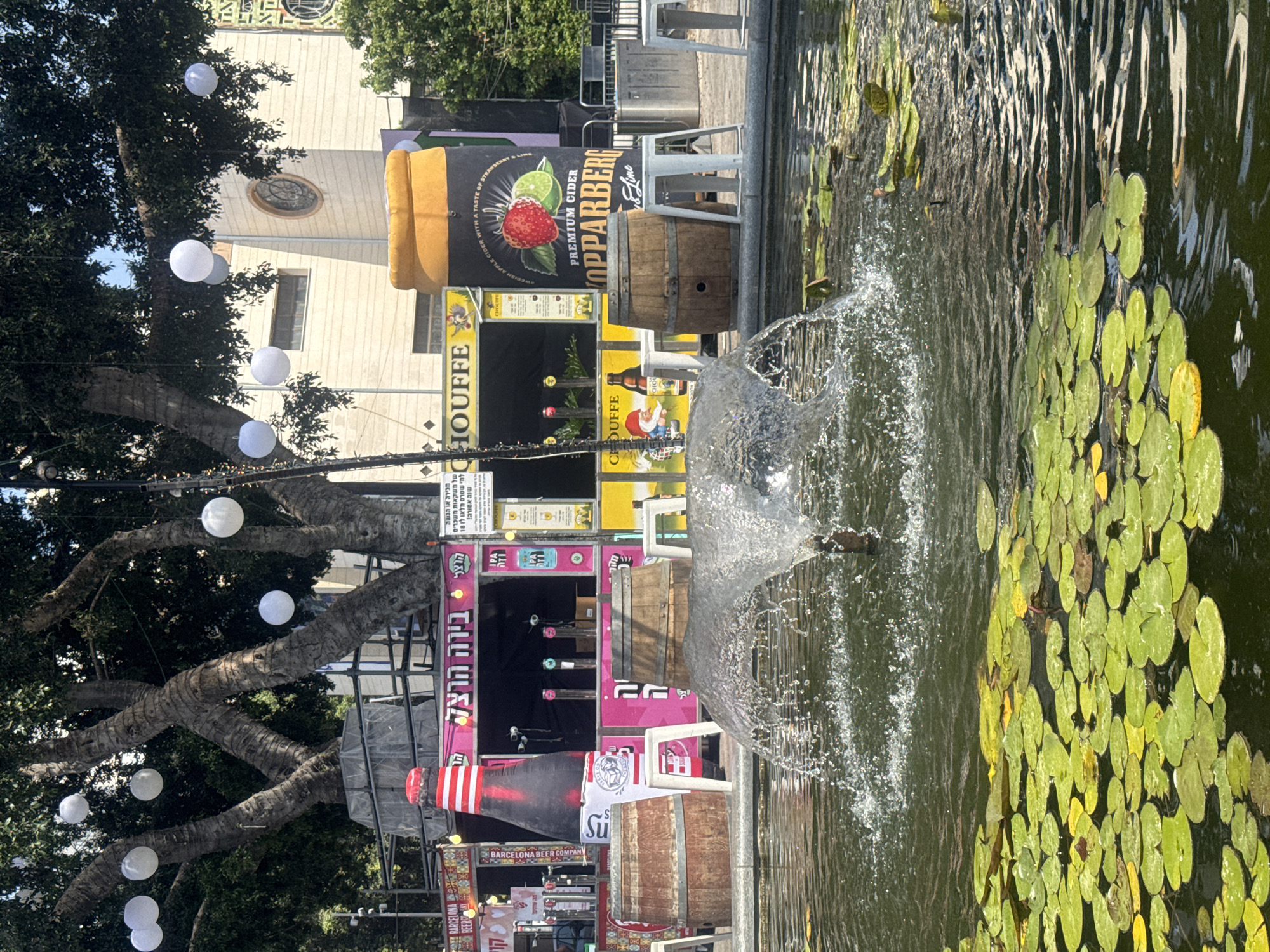The image depicts an outdoor scene in Ramat Gan, Israel, likely a public square or park. In the foreground, a water fountain is active, creating a spray of water. The fountain is surrounded by lily pads floating on the surface of a body of water. In the mid-ground, there are several market stalls or kiosks. One prominent stall features a large inflatable bottle and is decorated with pink and white colors. Another stall displays a large inflatable can for "Kopparberg Premium Cider" which shows a strawberry and lime. Wooden barrels are used as props or seating around the stalls. White plastic stools are also visible. In the background, a large tree with multiple trunks is adorned with white spherical lanterns hanging from its branches. Behind the tree and stalls, a building with an arched window can be seen. The overall atmosphere suggests a festive or recreational setting, possibly during a market event or fair.