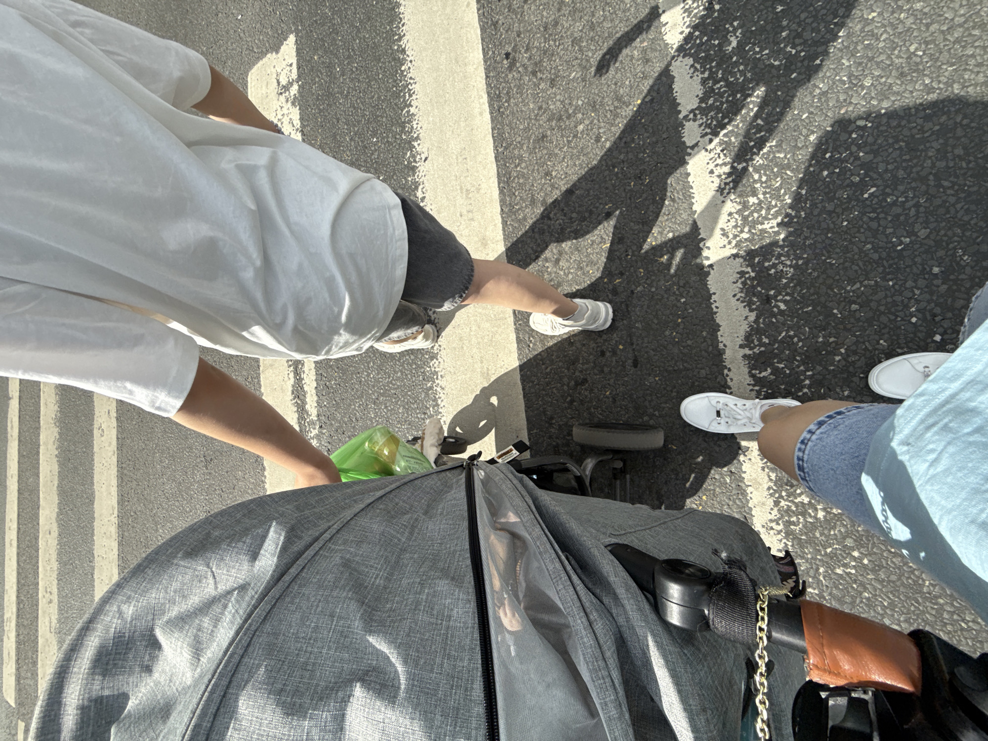 The image shows a close-up, overhead view of people walking across a pedestrian crossing in what appears to be a city. The perspective suggests the photo is taken from within a stroller or baby carriage. In the foreground, the grey fabric of a stroller is prominent, with a portion of its handle and a gold chain visible.
To the left, a person wearing a white t-shirt and dark shorts is seen from the waist down, with their legs and feet on the asphalt. They are wearing white sneakers and appear to be walking. A green plastic bag is also visible near their feet.
To the right, another person is partially visible, wearing blue denim shorts and white sneakers. Their legs and feet are on the pedestrian crossing.
The setting is an urban street with a clearly marked pedestrian crossing. The sunlight casts long shadows across the road, indicating it is likely daytime, possibly late afternoon or early morning. The ground is asphalt, and the pedestrian crossing stripes are white. The overall impression is of an everyday outing, likely a family walking with a stroller. No text is visible in the image.