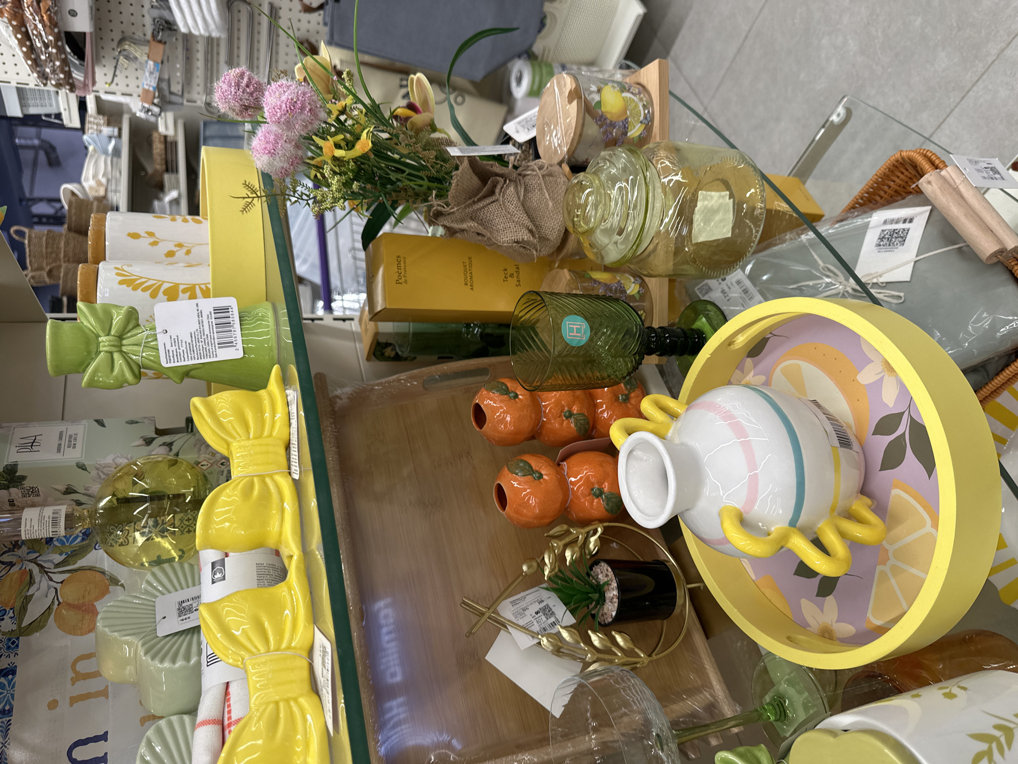 The image shows a retail display shelf in a store, likely a home decor or gift shop, featuring an assortment of decorative items. The shelf is made of glass and is supported by metal legs. On the shelf, there are several items including yellow bow-shaped decorations, orange ceramic vases shaped like tangerines, a green glass goblet, a clear glass jar with a wooden lid, and a bouquet of pink and yellow artificial flowers. In the foreground, a yellow tray holds a white vase with blue and pink stripes and yellow handles. Behind these items, there are more shelves with various products, including baskets, candles, and other decorative objects. The overall impression is one of a colorful and inviting display. There is no specific indication of the city of Samara, Russia, or any people in the image.