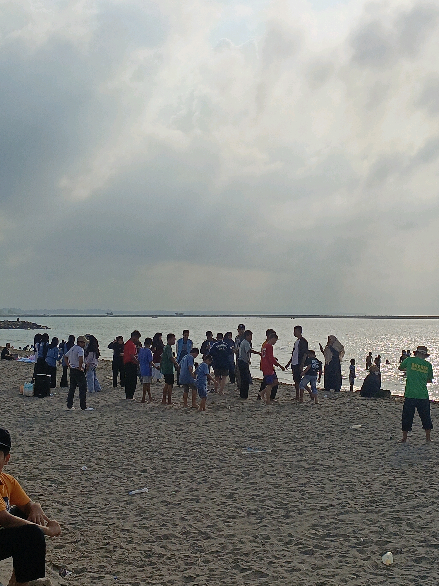 The image depicts a crowded beach scene on a cloudy day, likely in Bulukumba, Indonesia, given the context. The beach is covered in sand with some litter visible. A large group of people, comprising adults and children, are gathered on the sand, some standing and interacting, others walking. Several individuals are visible in the shallow water, with more people further out in the sea. The water is calm and reflects the overcast sky. In the background, a distant shoreline with some structures is visible, and further out at sea, two boats or small vessels can be seen. The overall atmosphere suggests a casual outing or recreational activity at the beach.