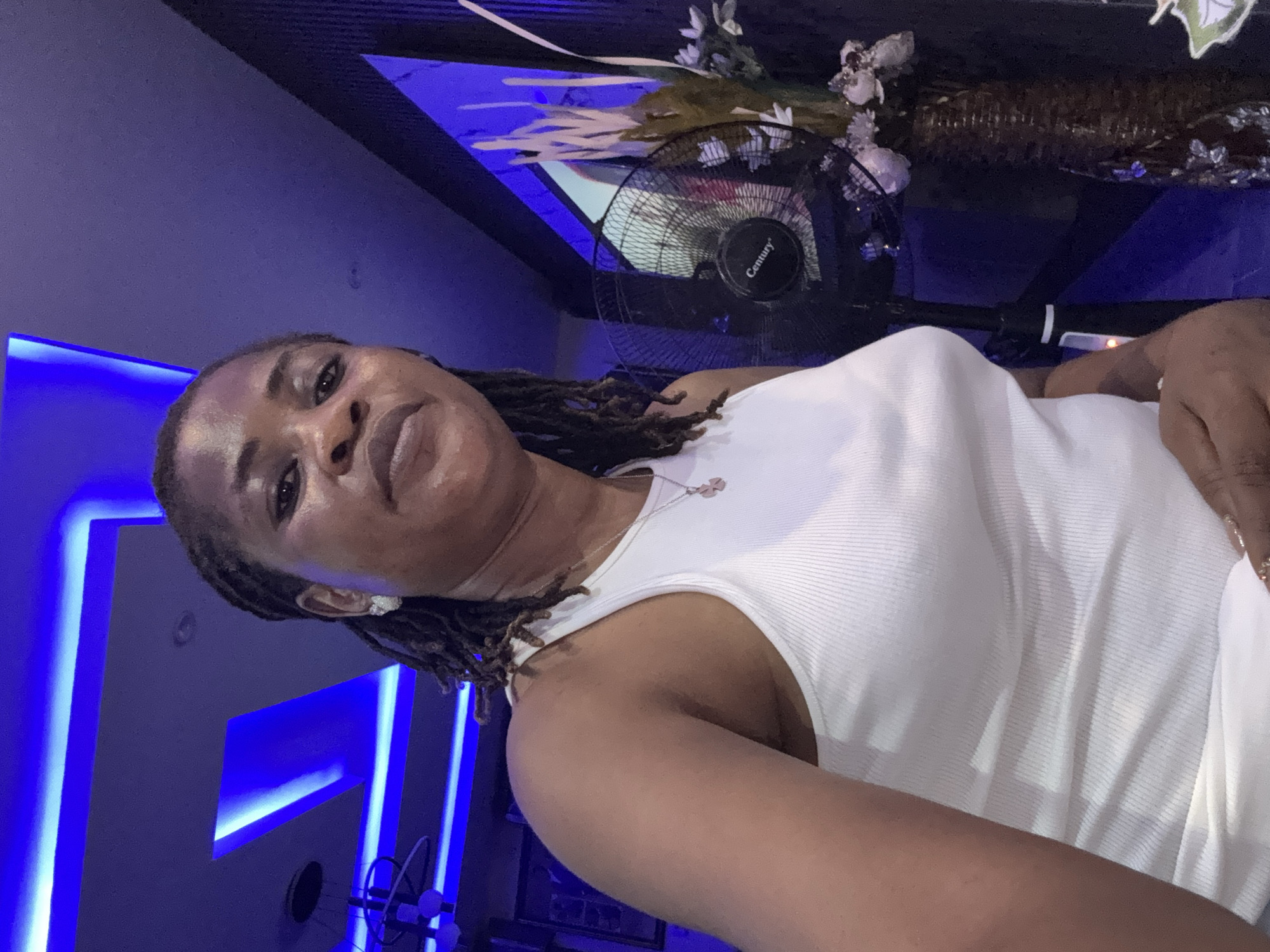 A medium shot captures a woman with dreadlocks wearing a white ribbed tank top, looking directly at the camera in a room with blue ambient lighting. She has subtle makeup on her eyes and lips, and she is wearing a necklace with a small pendant and earrings. To her right, a black oscillating fan with a "Century" logo is visible, and behind it, a decorative vase with dried flowers and some shimmering embellishments stands on a dark surface. The background features a wall with vertical paneling and a framed picture. The ceiling has recessed lighting with blue neon strips. The overall scene suggests an indoor setting, possibly a lounge or a styled room, in Ajara, Nigeria.
