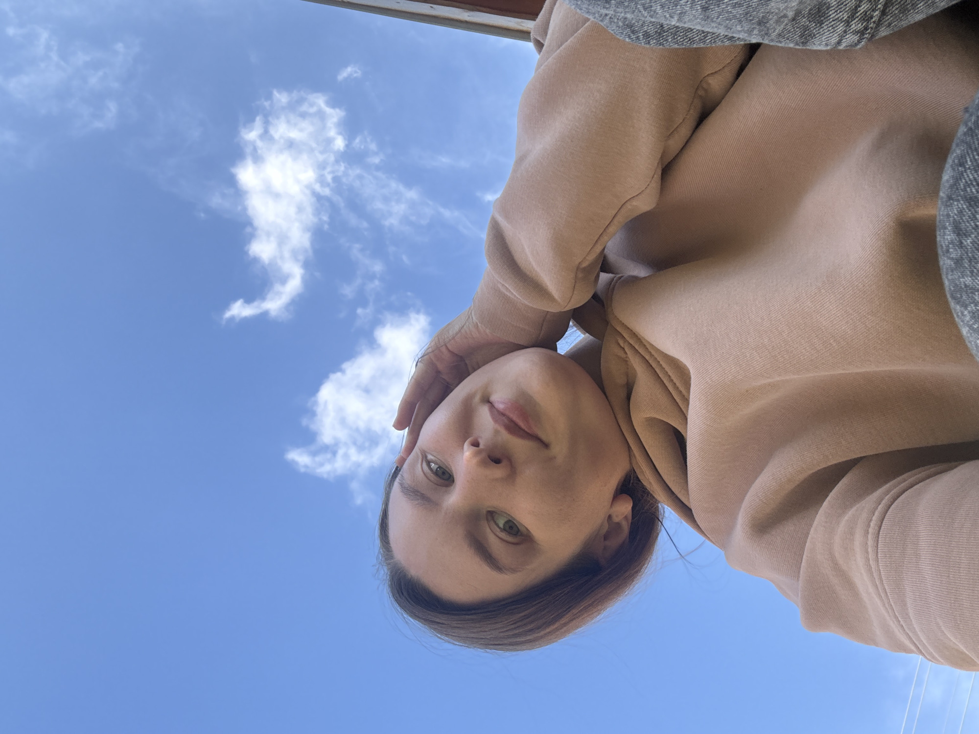 A low-angle selfie shows a person with light skin, green eyes, and light brown hair, wearing a beige hoodie and denim clothing, likely taken outdoors under a blue sky with scattered white clouds. The location is unidentifiable as Italmas, Russia, without more information in the image.