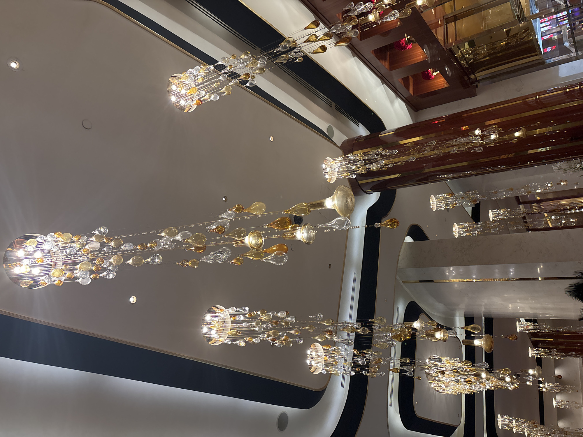 The image captures a wide-angle view of an interior space with ornate chandeliers. The chandeliers are made of numerous crystal-like elements in clear, amber, and gold hues, suspended from thin wires. They are arranged in a cascading fashion, creating a dazzling effect. The ceiling is a neutral color, likely white or light grey, with some subtle architectural details like curved lines and dark trim. A few recessed lights are visible on the ceiling.
The scene suggests a luxurious and possibly grand setting, such as a hotel lobby, a ballroom, or an upscale shopping mall. The lighting from the chandeliers creates a warm and inviting ambiance. There is a sense of depth and scale, with multiple chandeliers visible at different distances and levels. The reflections on some of the surfaces hint at the polished nature of the interior design. No people are visible in the image. The image does not contain any visible text. The time of day is indeterminate due to the indoor setting. The overall impression is one of elegance and sophistication.