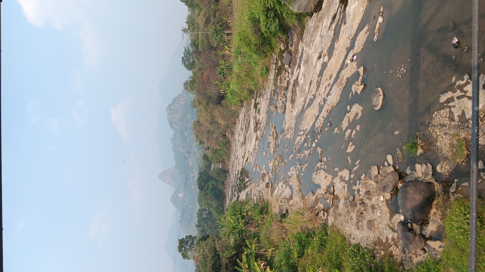 This is a vertical, eye-level photograph of a shallow, rocky riverbed with water flowing through it. The river is situated in a rural area with lush green vegetation on either side, including trees, banana plants, and grass. In the distance, there are rolling hills and mountains, shrouded in a light haze. The sky is a pale blue with a few scattered clouds. The foreground shows a black railing, suggesting the photo was taken from a bridge or elevated vantage point. The riverbed is characterized by large, flat rocks and smaller stones, with patches of exposed sand and sediment. There is some visible litter in the river, including plastic bags and debris. A bird is visible in the sky, flying high above the landscape. The overall impression is one of a natural, somewhat untamed landscape.