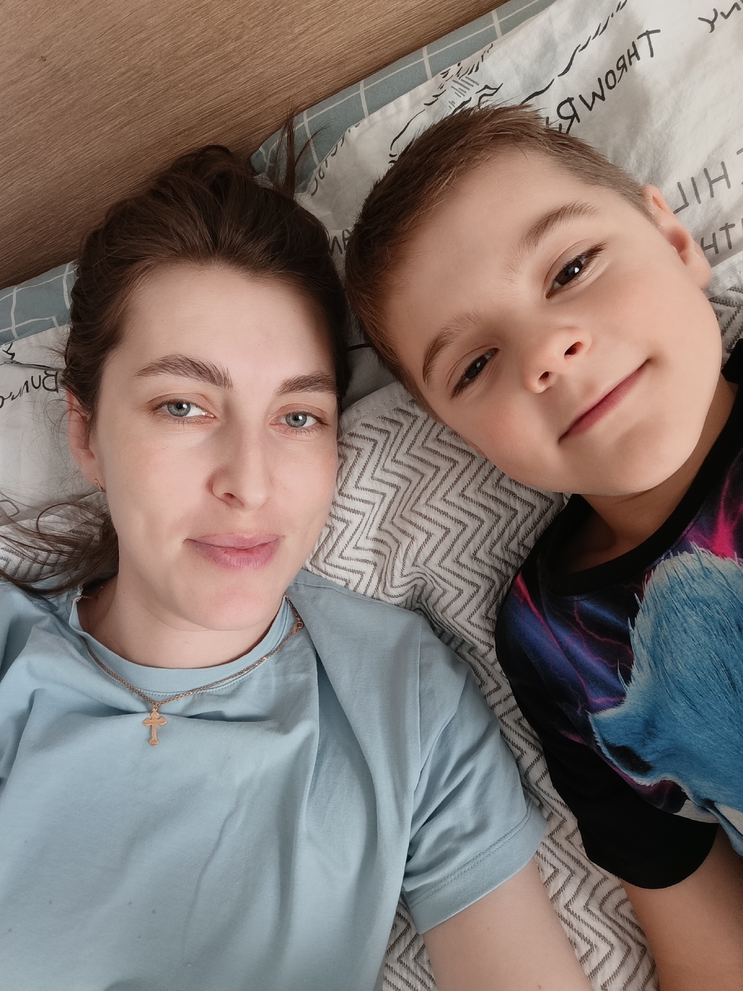 A woman and a young boy are lying next to each other on a bed. The woman has brown hair pulled back, blue eyes, and is wearing a light blue t-shirt with a gold cross necklace. The boy has short brown hair and is wearing a black t-shirt with a blue and purple design. They are both looking at the camera. The bed has patterned sheets and pillows. The location is Chaikovsky, Russia.