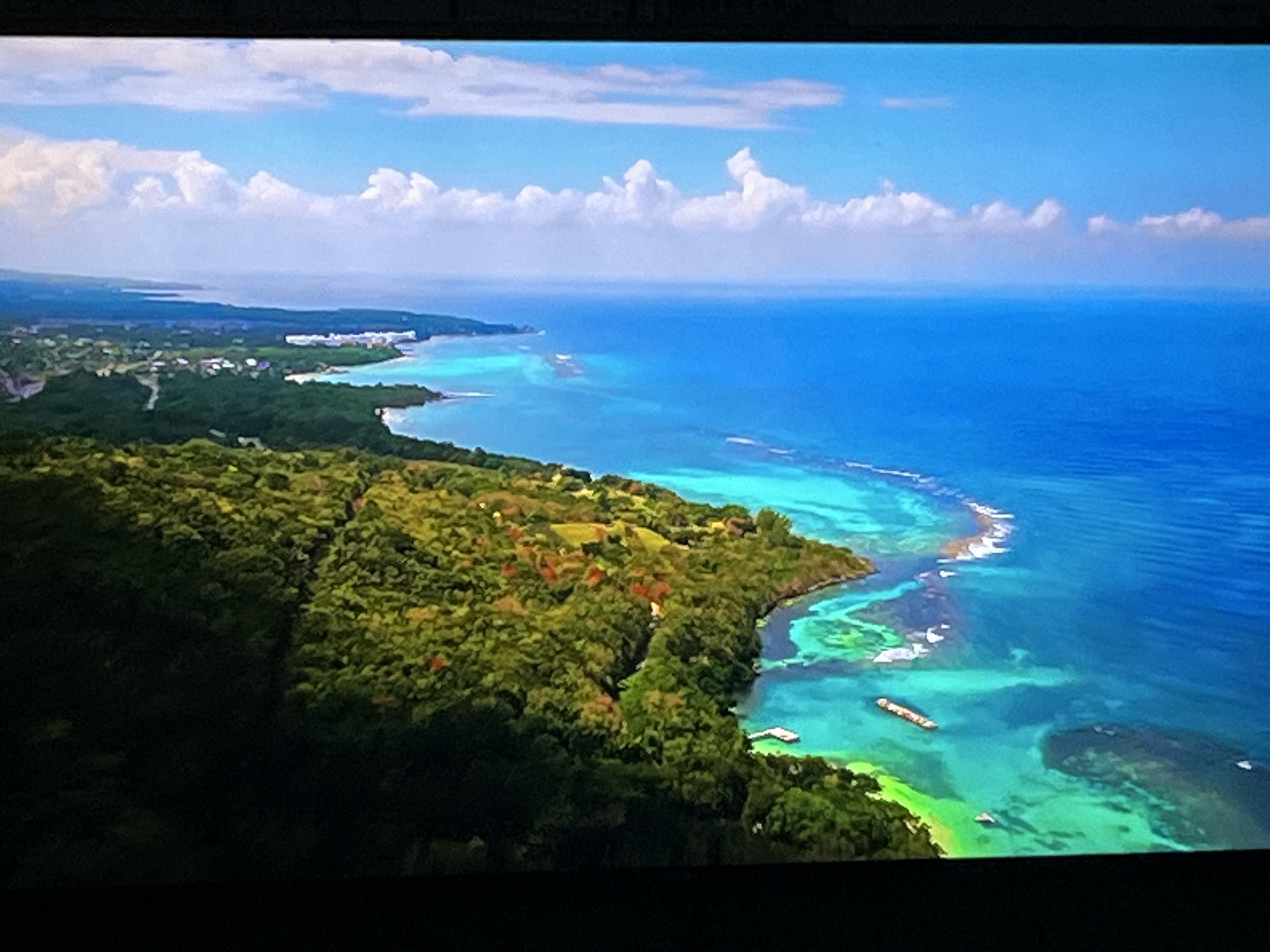 This image is an aerial view of a lush, green coastline meeting a vibrant blue ocean. The land is densely forested, with hints of red roofs visible amongst the trees, suggesting scattered settlements or villas. The ocean is clear and turquoise near the shore, revealing coral reefs and shallow areas before deepening into a rich blue further out. Several boats, including what appears to be a barge, are visible on the water. In the distance, the coastline stretches out, with more buildings and greenery along the shore. The sky is a bright blue, dotted with fluffy white clouds. The scene is serene and picturesque, depicting a tropical or subtropical coastal landscape.
The provided image and crops show no recognizable landmarks or features that would allow for identification of the city of Kharkiv, Ukraine. The environment depicted is consistent with a tropical or subtropical coastal region, which is not characteristic of Kharkiv.