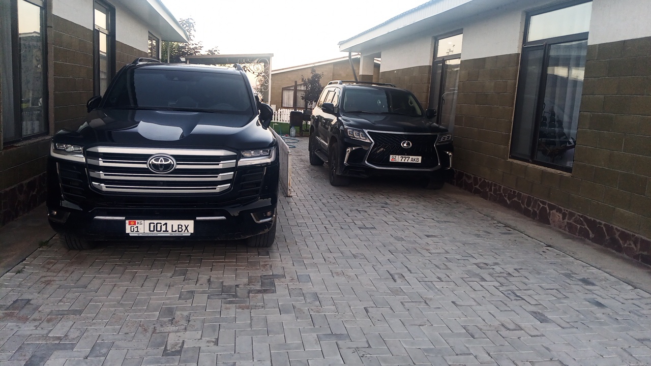 In the city of Choktal, Kyrgyzstan, two black SUVs are parked side-by-side on a paved driveway. The vehicle in the foreground, a Toyota Land Cruiser, has a license plate displaying "KG 01 001 LBX". To its right, a Lexus SUV has a license plate that reads "02 777 AKB". The SUVs are positioned in front of a building with brick-patterned walls and large windows, suggesting a residential or commercial setting. The ground is covered with grey brick pavers arranged in a herringbone pattern. The scene appears to be outdoors, with a bright sky visible, indicating daytime. No people are present in the image.