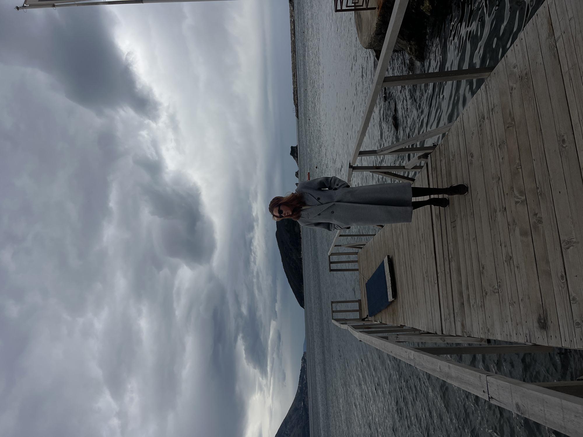 A woman in a gray coat is standing on a wooden pier with a ladder in Budva, Montenegro. The sky is overcast with dark clouds. Water surrounds the pier. A mountain is visible in the background.