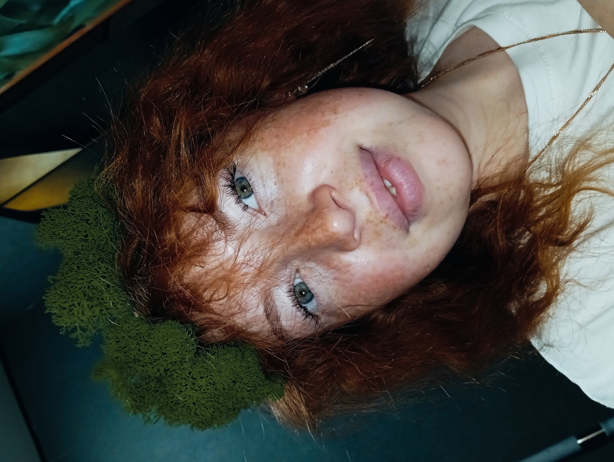 A close-up, tilted portrait shows a young person with striking red hair, a prominent feature in the image. Numerous freckles are visible across their nose and cheeks, and their eyes are a clear light blue or green, looking directly upwards towards the camera. Their lips are slightly parted. They are wearing a simple white top and a delicate gold chain necklace.
To the left of their head, a dense, irregular mass of green, moss-like material rests, providing a contrasting texture and color against the person's red hair and fair skin. In the hair, a small, metallic, braided hair accessory can be seen. The background is dark and largely out of focus, suggesting an indoor setting. Some faint, abstract shapes or patterns, possibly part of furniture or decor, are discernible in the upper left corner. The lighting appears soft, highlighting the features of the person. The overall impression is a natural and intimate pose. No specific activity beyond posing for the photograph is depicted, and no text is visible. The image is set in Yurgamysh, Russia.