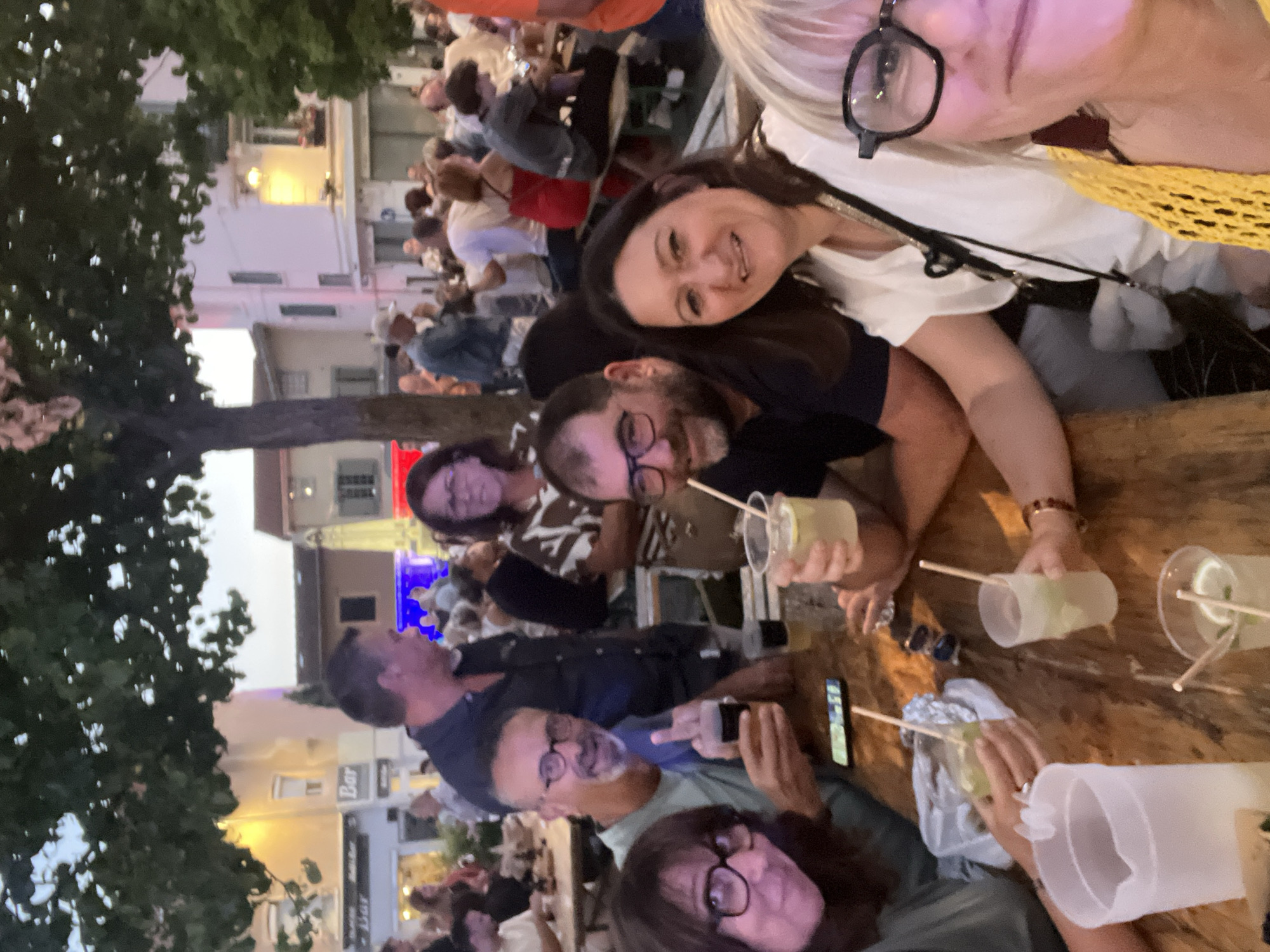 This is a candid, medium-distance, outdoor photograph taken at ground level on a wooden table at what appears to be an outdoor bar or restaurant in Saint-Galmier, France. The lighting is low, suggesting it is late afternoon or early evening. A group of people are gathered around the table, some holding drinks. In the foreground, a woman with blonde hair and dark-rimmed glasses is partially visible from the chest up, wearing a yellow knitted top. To her left, a woman with dark hair and glasses is smiling towards the camera. To her left, a man with glasses and a beard is holding a dark drink and smiling. Further left, another woman with glasses is visible. In the mid-ground, more people are seated at the table and in the background, the scene opens up to an outdoor gathering with buildings and trees. A "Bar" sign is visible in the background on a building.