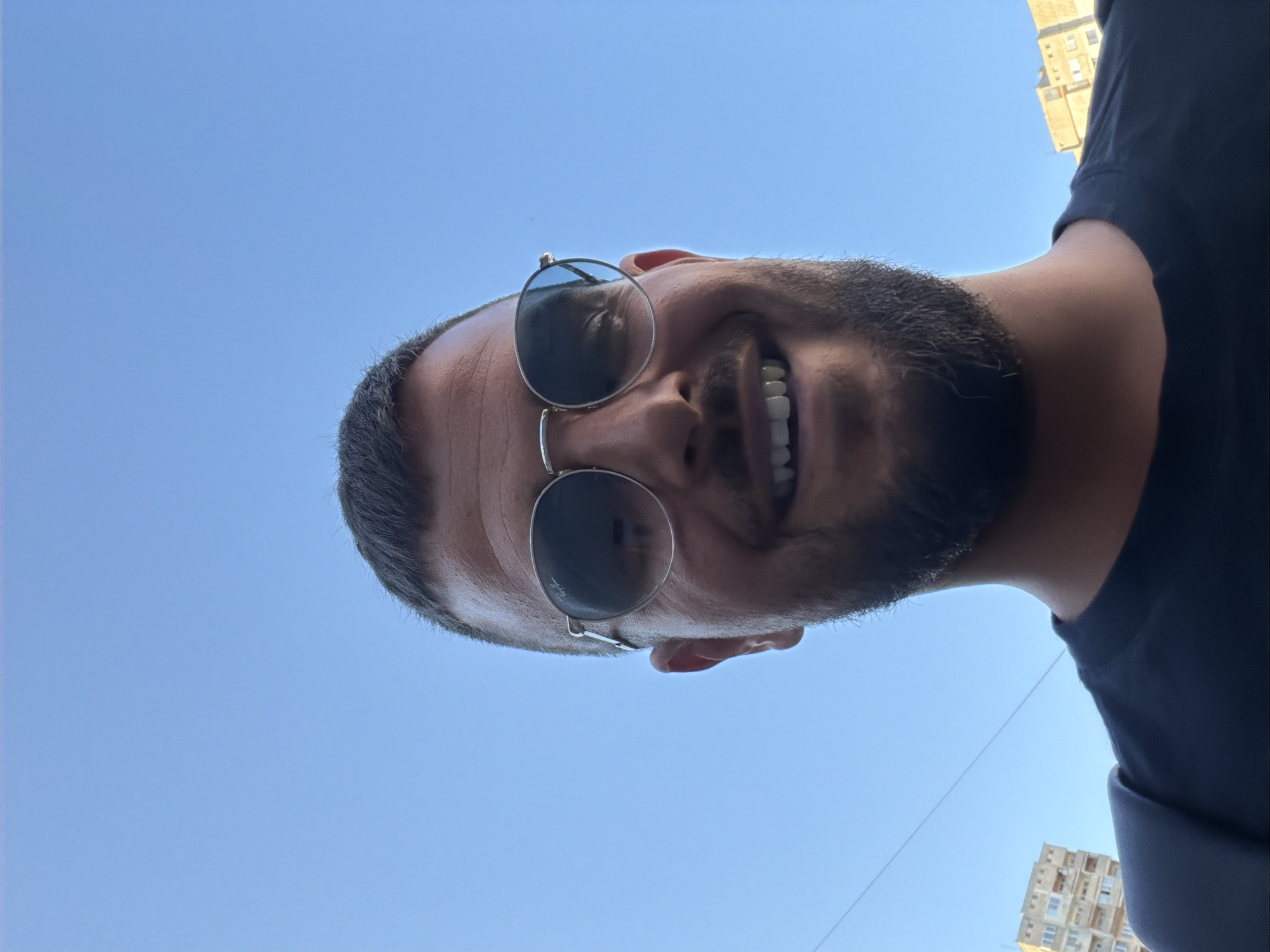 A man wearing sunglasses and a black t-shirt stands outdoors with a clear blue sky behind him. Buildings are visible in the background. The image is a selfie, taken from a low angle, looking up at the man's face. He has a beard and is smiling. The location is the city of Sofia, Bulgaria.