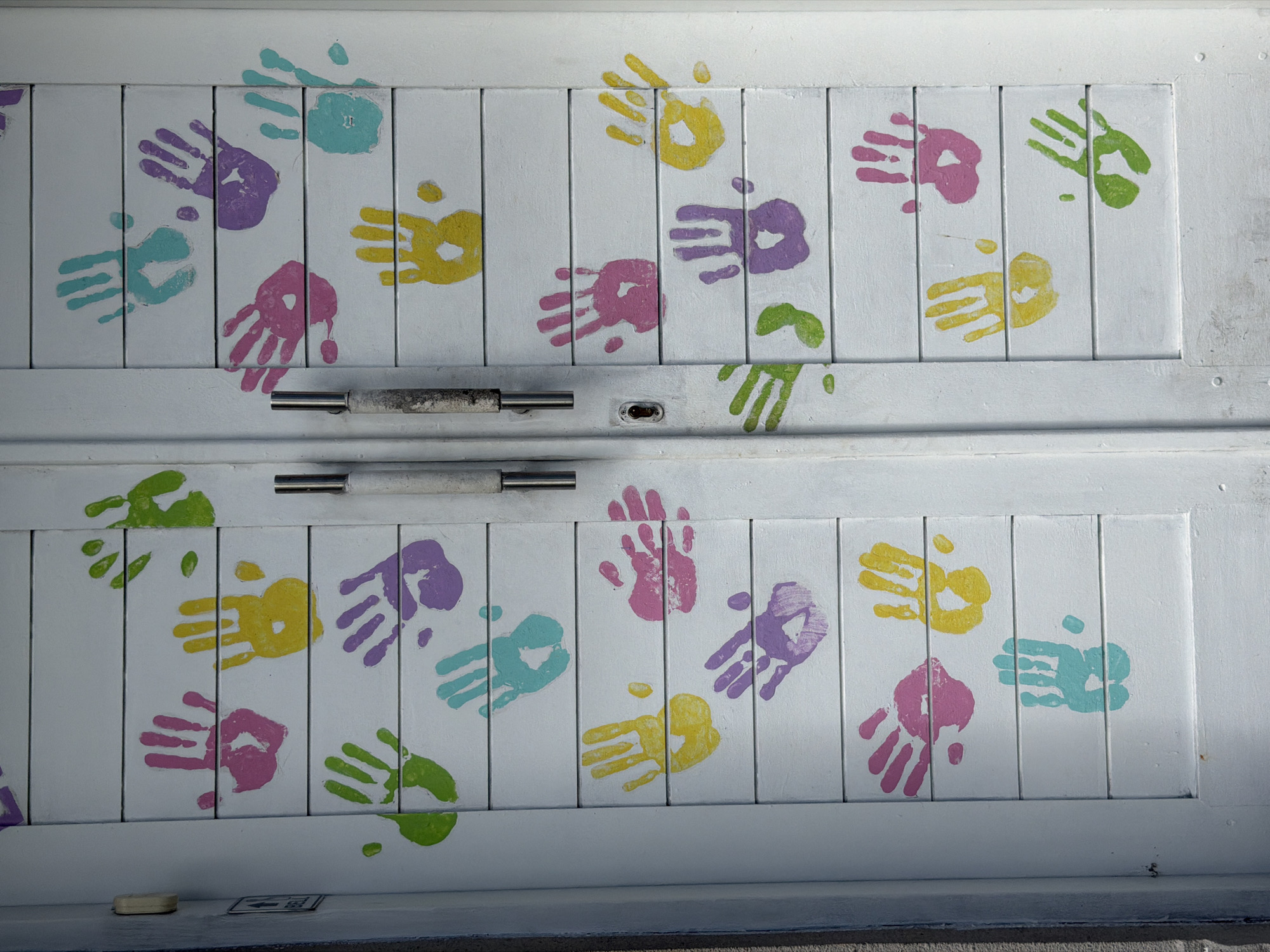 A close-up, full-shot photograph depicts a set of closed white-painted wooden double doors adorned with multicolored handprints. The handprints are in various colors including purple, yellow, pink, green, and teal. The doors appear to be made up of vertical wooden panels. A metal handle spans the center of the doors, separating the top set from the bottom set. The doors appear to be part of a building or structure. The photograph was taken during daylight hours, presumably in Kuta, Indonesia. The image contains no people.