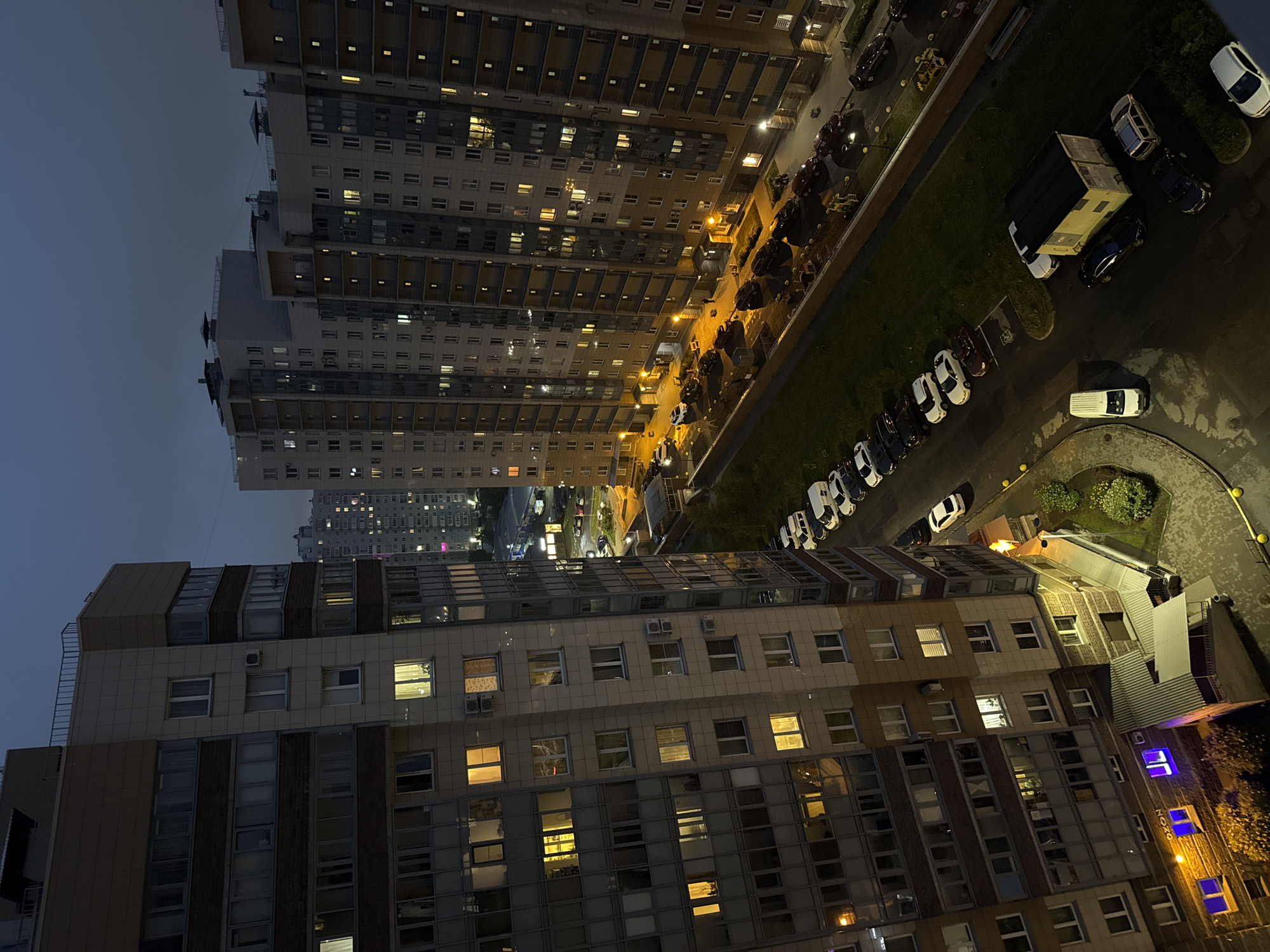 The image depicts a view from a high vantage point overlooking a residential area at dusk. Two tall, modern apartment buildings, constructed with a combination of beige, brick, and glass facades, dominate the foreground and midground. The building on the left is closer and more detailed, revealing numerous windows, some illuminated from within, suggesting occupancy. The building on the right is further away, with its illuminated windows creating a patterned effect against the darkening sky.
Below the buildings, a paved area serves as a parking lot and access road. Several cars of various colors and models are parked along the side of the road and in designated spots. The vehicles appear to be stationary. A few scattered trees and patches of grass provide some greenery in the urban landscape. In the distance, more apartment buildings are visible, their lights creating a diffused glow. The sky is a deep, dusky blue, indicating either early evening or late twilight. The overall scene conveys a sense of a typical urban residential environment after sunset.