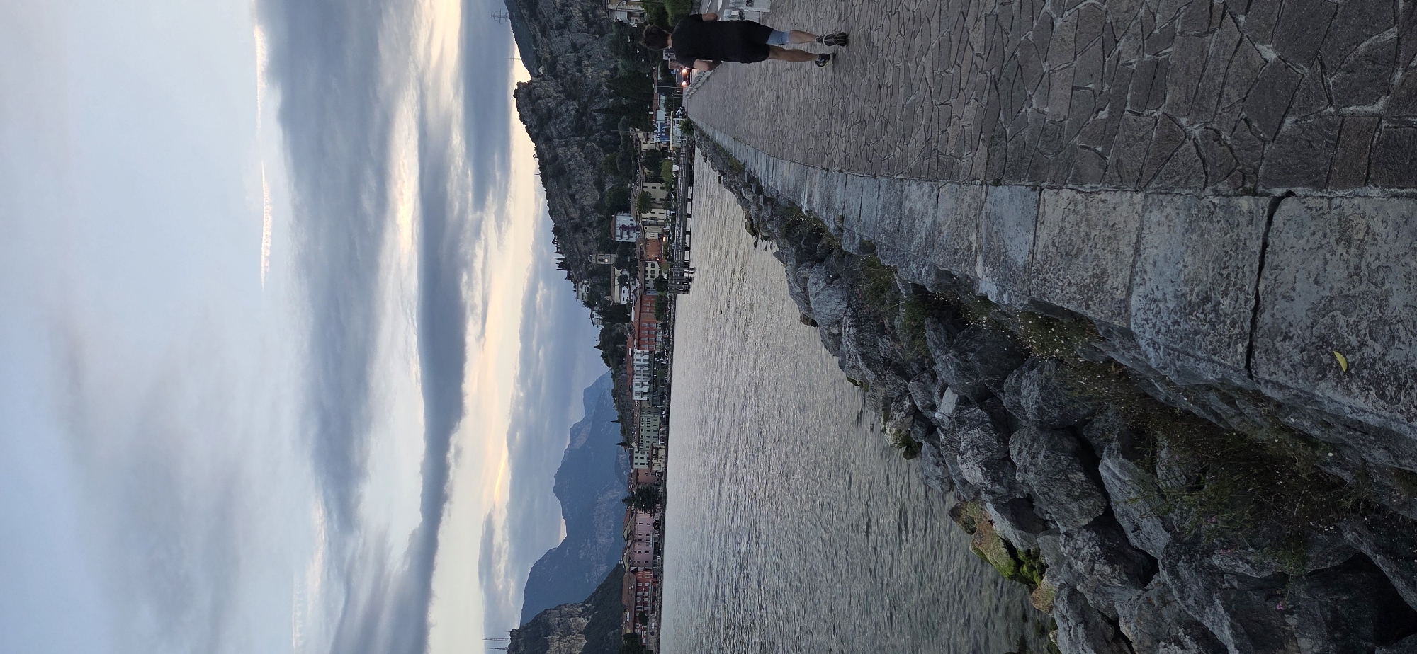 The image depicts a scenic view of the waterfront promenade in Torbole, Italy, during twilight. A lone figure, a male with dark hair wearing a black t-shirt and shorts, is jogging away from the camera on a paved walkway. The walkway is lined with large, grey rocks and overlooks a body of water, likely Lake Garda. In the background, a picturesque town with colorful buildings is nestled against a backdrop of dramatic, mountainous terrain under a cloudy sky. The overall atmosphere is serene and hints at the end of a day with subdued lighting.