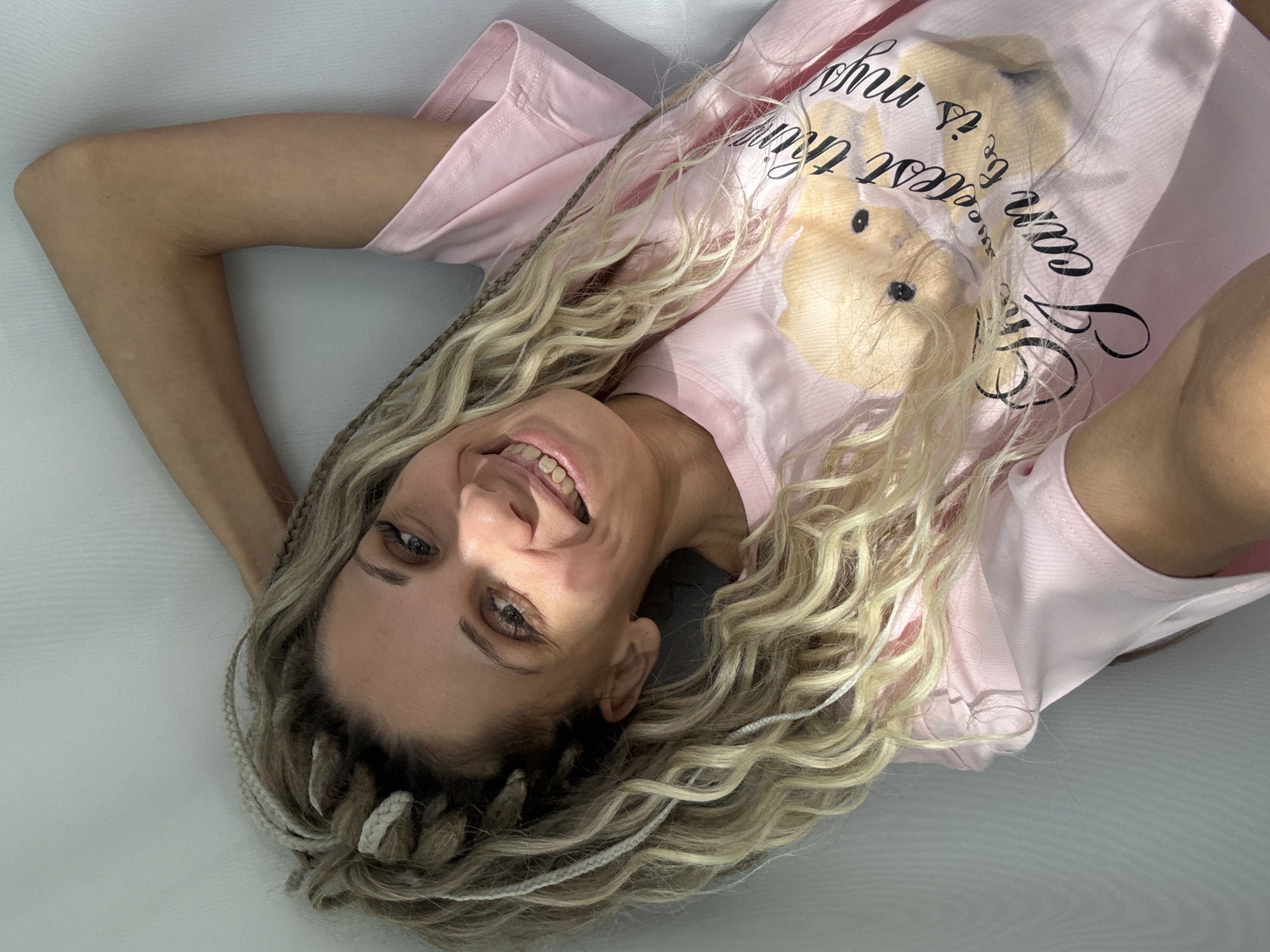 A woman lies on a white surface, possibly a bed or couch. She is wearing a light pink t-shirt with a cartoon bear print and black text. She has long, blonde, braided hair. Her left arm is bent and resting above her head, while her right arm is extended to the right. She is smiling and looking at the camera. The background is a plain white surface. There is no discernible evidence to support a location in Rusanovo, Russia.