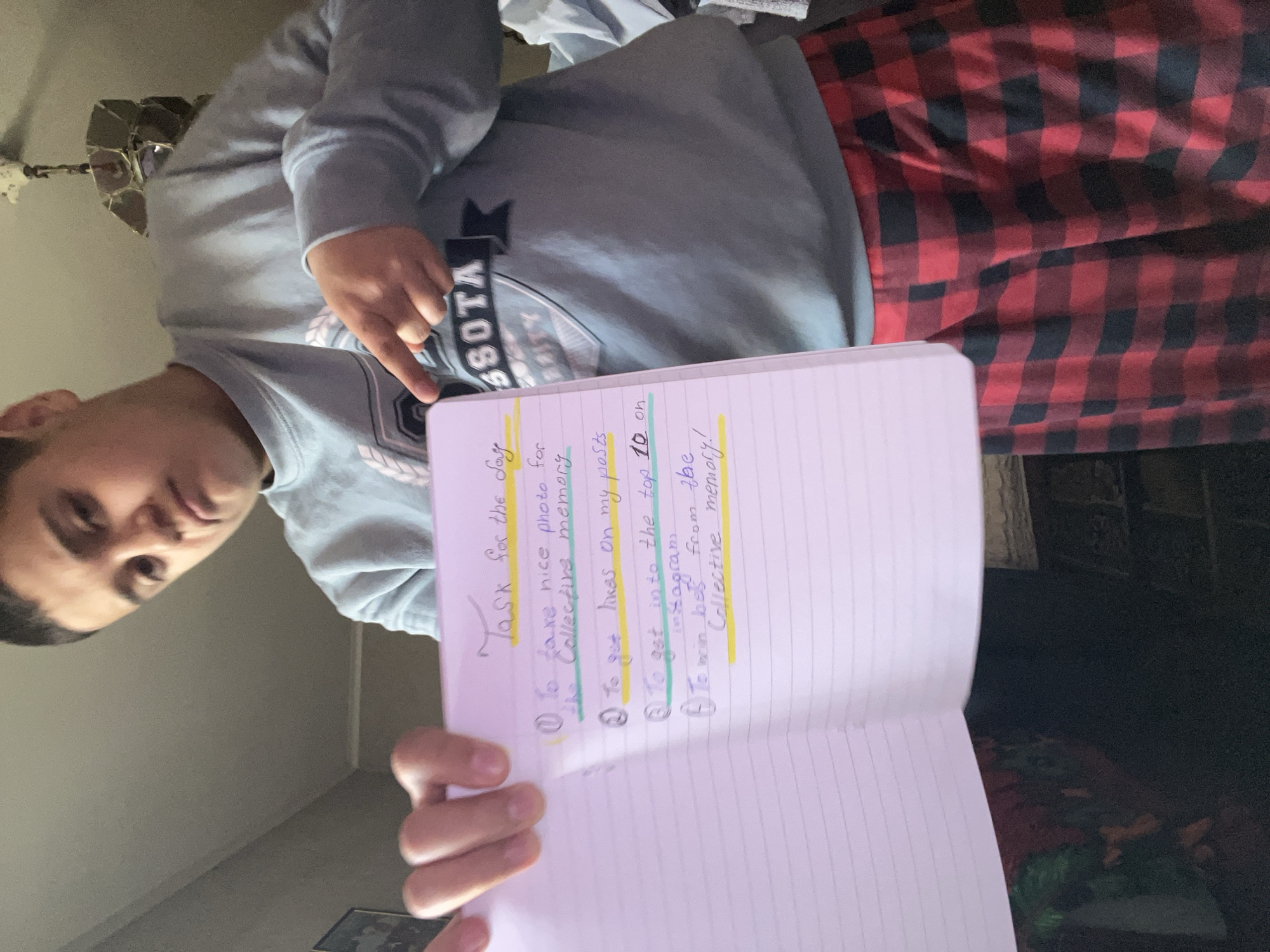 An indoor, low-angle shot in Lozarevo, Bulgaria, shows a young person, likely male, holding an open notebook toward the camera. The person, positioned in the upper left of the frame, wears a light blue sweatshirt with "SOTA" (likely part of "MINNESOTA") and a crest design visible, paired with red and black plaid pajama bottoms. They are pointing a finger, possibly at the notebook or gesturing. Their expression is neutral as they look directly at the viewer.
The notebook, held prominently, displays handwritten text in various colored inks (purple, blue, green) and highlighted sections (yellow, green, blue). The title reads "Task for the day." The listed tasks include:
1. "To take nice photo for the Collective memory"
2. "To get likes on my posts."
3. "To get into the top 10 on instagram"
4. "To win BIG from the Collective memory!"
The background is dimly lit, revealing a light-colored wall, a partially visible, decorative ceiling fan in the upper left, and a blurry, dark area to the right, possibly containing furniture. The casual attire and indoor setting suggest a relaxed environment, possibly a home.
