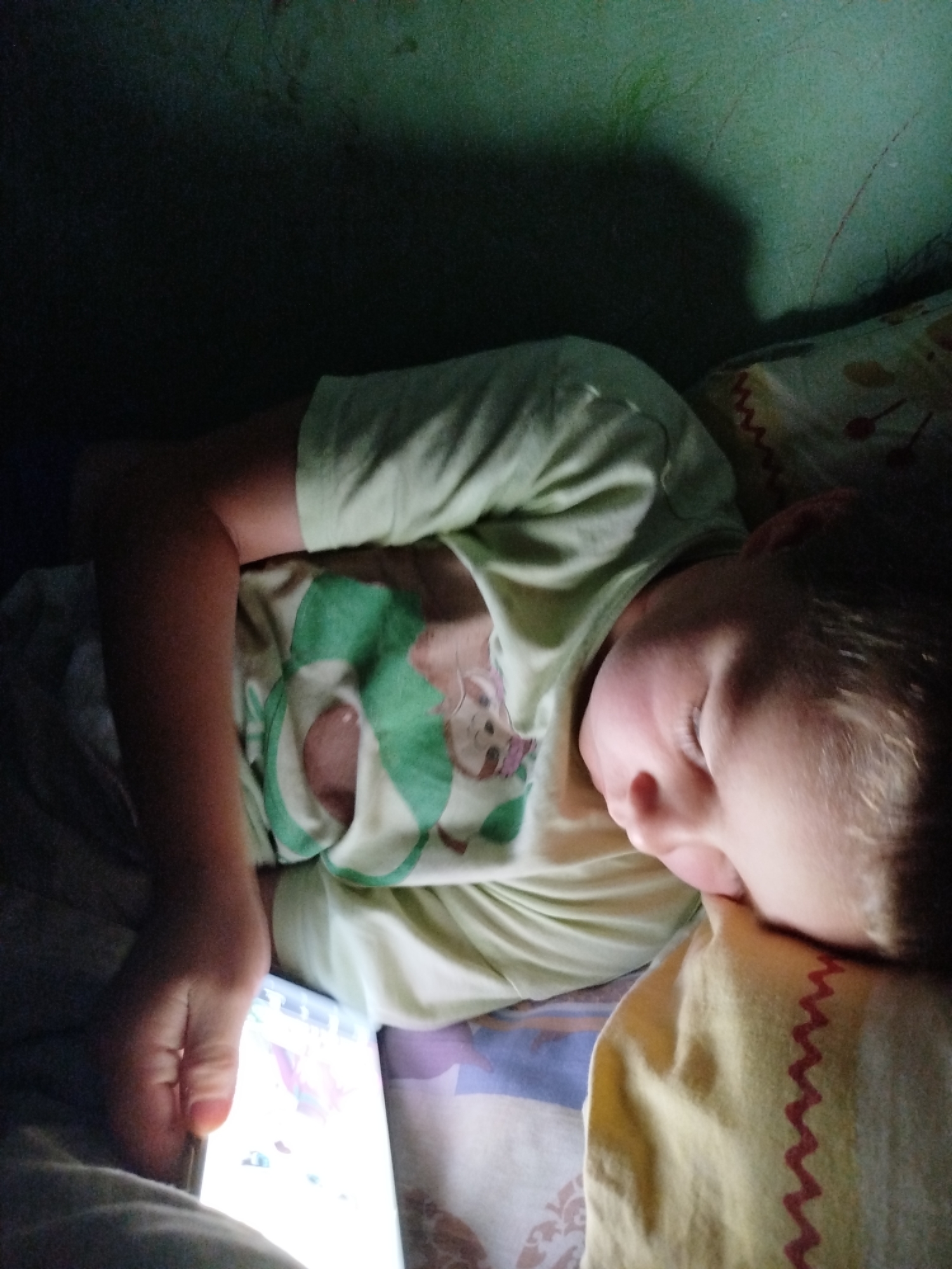 The image shows a close-up of a child sleeping, holding a phone. The child is lying on a bed with a light green pillow and a yellow pillow with red zig-zag stitching. The child is wearing a light green t-shirt with a cartoon image on it. The room appears to be dimly lit, with shadows cast on the wall. The location in the city of Крывасельскі сельскі Савет, Belarus is not determinable from the image alone.