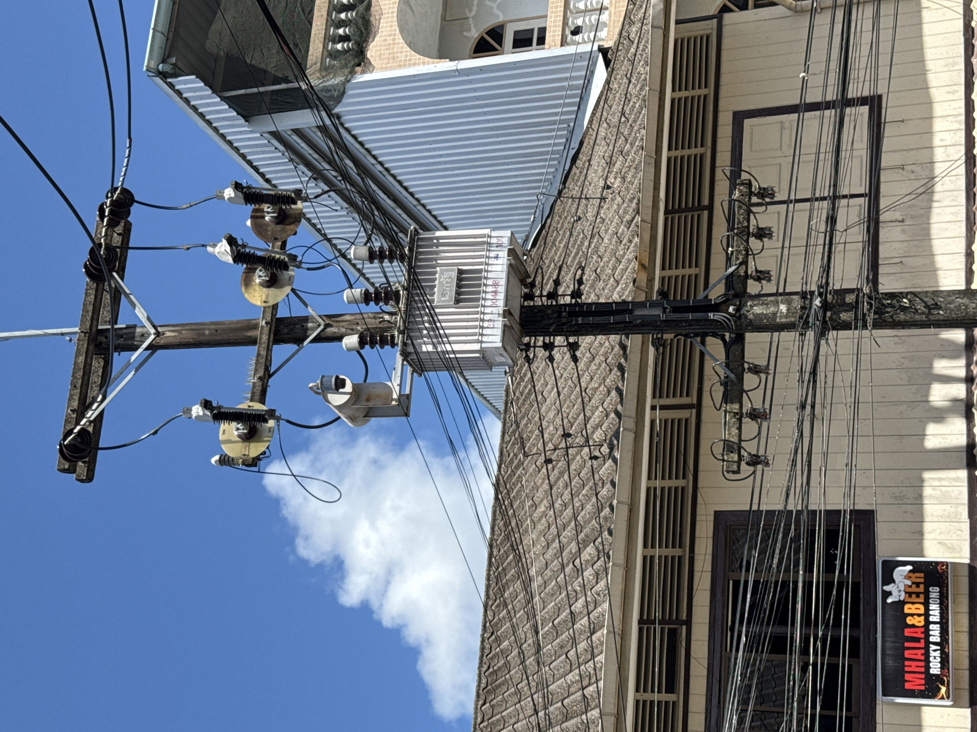 The image shows a utility pole with electrical transformers and wires in the city of Ranong, Thailand. A sign at the bottom left reads "MHALL&BEER ROCKY BAR RANONG". The pole is situated in front of buildings with tiled roofs and paneled walls. The sky is blue with some white clouds. There are no people visible in the image.