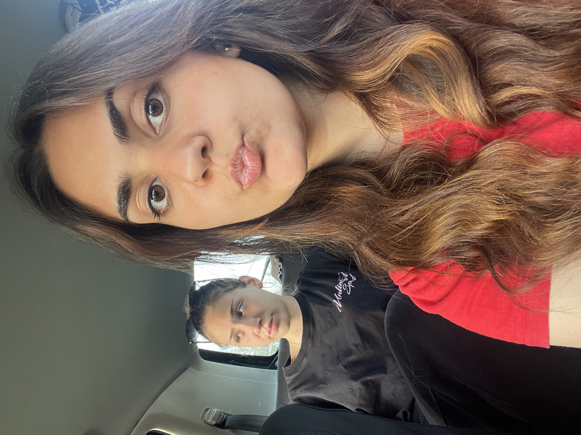 The image depicts two young women in what appears to be the interior of a car in the city of Unknown, Unknown. The primary subject is the woman in the foreground, who is fair-skinned, with brown hair and brown eyes. She is wearing a red top, and has her hair in loose waves with blonde highlights. She is making a "kiss" face for the camera.
In the background, a second woman is visible. She has fair skin, dark hair pulled back and is wearing a black t-shirt with a white logo. She is sitting in what appears to be the backseat and is looking at the camera. The car's interior is visible with gray panels and seat covers. The photo appears to be taken during the daytime, with natural lighting visible. The setting is suggestive of a routine, everyday scenario of driving.