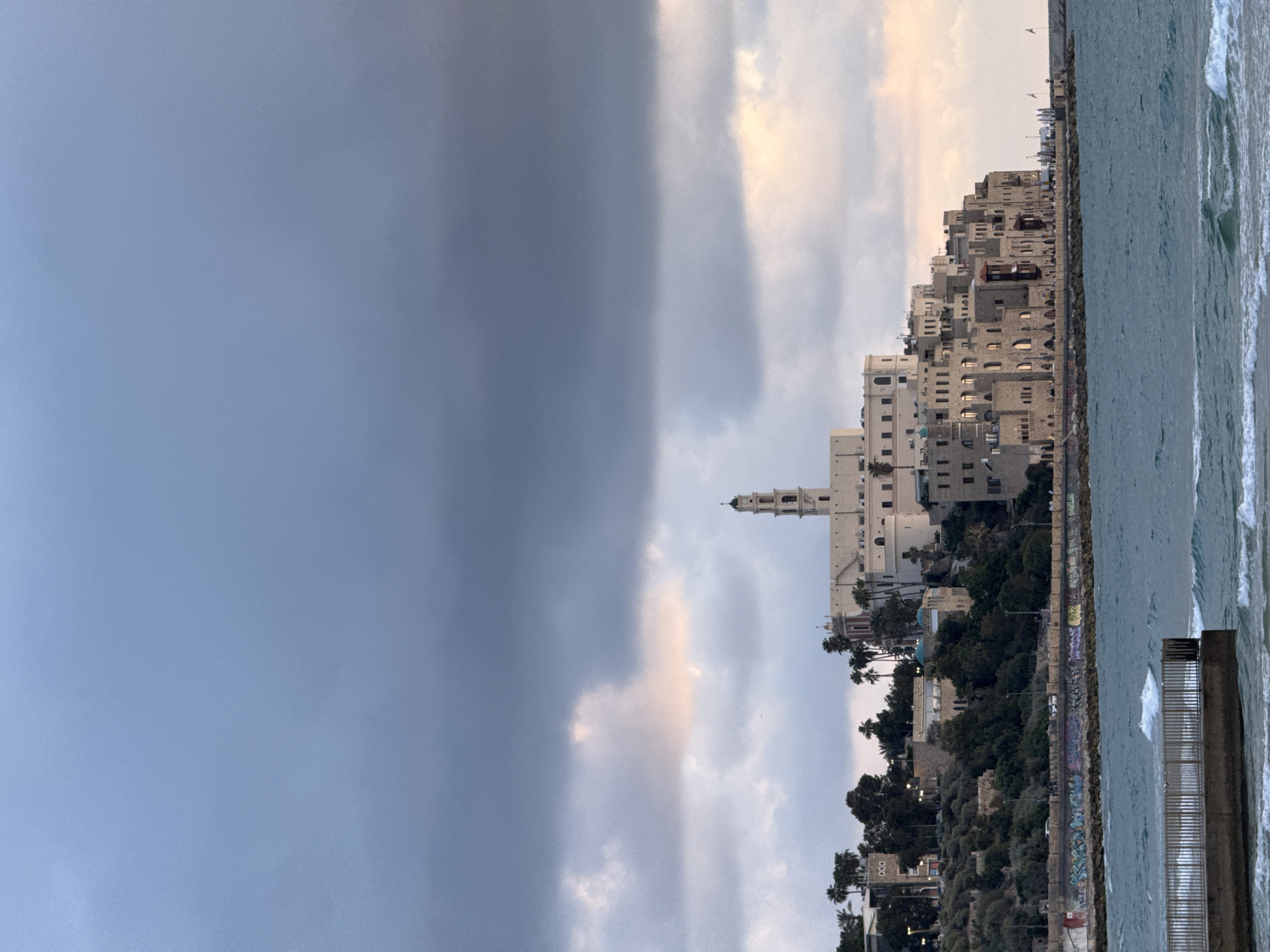 The image depicts the Mediterranean coastline of Tel Aviv, Israel, with a contrasting sky divided between a dark storm cloud and a lighter, cloud-filled expanse. Dominating the center is the old city of Jaffa, characterized by its historic architecture, including a prominent church tower. Lush greenery and trees cover the area below the buildings, leading down to a seawall covered in graffiti and the turbulent ocean. The horizon line is defined by the meeting of the sky and the sea.