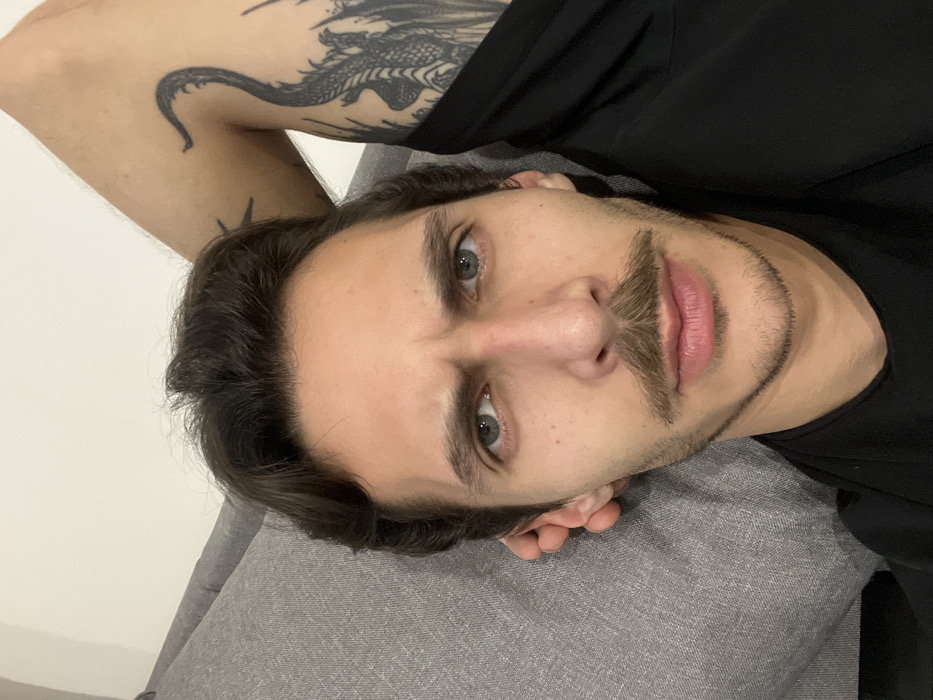 A man is lying on his back with his head tilted slightly towards the camera. He has light skin, brown hair, a mustache, and a light beard. His eyes are a pale green or blue. He is wearing a black shirt. His left arm is visible with a tattoo of a dragon on his upper arm. He is lying on a grey couch. The background appears to be a plain white wall.