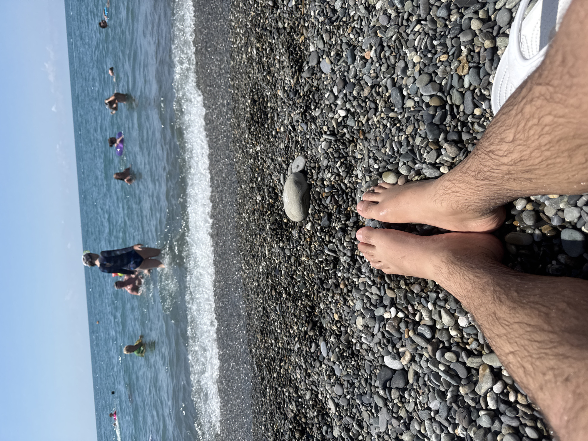 The image shows a pebble beach in Sochi, Russia, with several people swimming in the Black Sea under a clear blue sky. In the foreground, the legs and feet of an individual are visible, resting on the pebbles. Some of the swimmers are using inflatable rings. The water is calm with small waves approaching the shore.