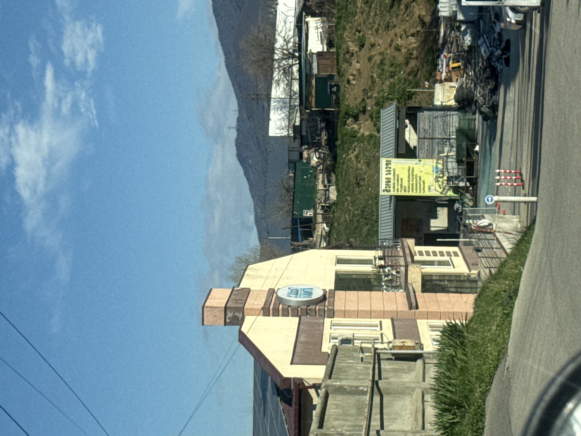 The image shows a view from a vehicle, possibly a car, in what appears to be a suburban or semi-urban setting. A building with a sign, likely a business or establishment, is situated on the left side of the road, and a mountain is visible in the background. There are powerlines crossing the blue sky. The building features a sign with possible English letters. The surrounding area includes vegetation and some other structures. The road is paved, with a strip of greenery alongside it, and a section of a vehicle's interior (possibly the dashboard or steering wheel) is visible at the very bottom. The overall impression is of a typical scene from the periphery of a city, possibly Tbilisi, Georgia.
