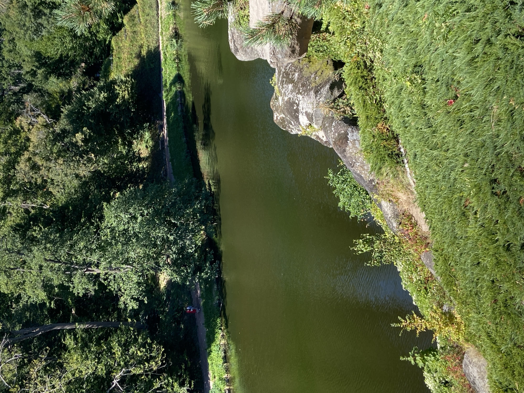 The image depicts a park setting with a body of water, likely a river or lake, in the foreground and mid-ground. Dense green trees and foliage line the banks of the water, with a pathway visible on the far bank. Two individuals, one in red and one in blue, are walking along this pathway. Large rocks and lush green ground cover are visible in the foreground, suggesting a landscaped area. The lighting indicates it is daytime, with sunlight casting shadows. The overall scene is serene and natural, with a focus on the interplay of water, vegetation, and geological features.