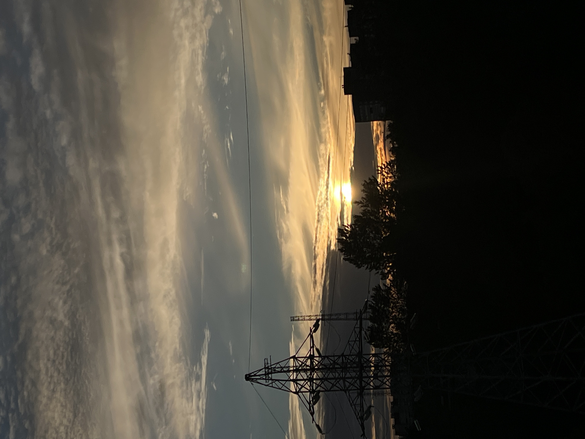 The image depicts a dramatic sunset over a cityscape, possibly Saint Petersburg, Russia. The sky is filled with wispy, elongated clouds catching the golden light of the setting sun, creating streaks of orange, yellow, and white against a pale blue background. The sun itself is a bright, diffused orb just above the horizon, casting a warm glow.
In the foreground and midground, silhouetted against the vibrant sky, are power transmission towers. One large, lattice-style tower dominates the lower left of the frame, its intricate metalwork starkly defined. Another, smaller tower is visible in the distance. The dark silhouettes of trees and buildings are also present on the right side of the image, suggesting an urban or semi-urban environment. The overall scene conveys a sense of natural beauty juxtaposed with industrial infrastructure. The time of day is clearly sunset due to the intense golden light and the position of the sun. The weather appears clear with scattered clouds. No text is visible in the image.