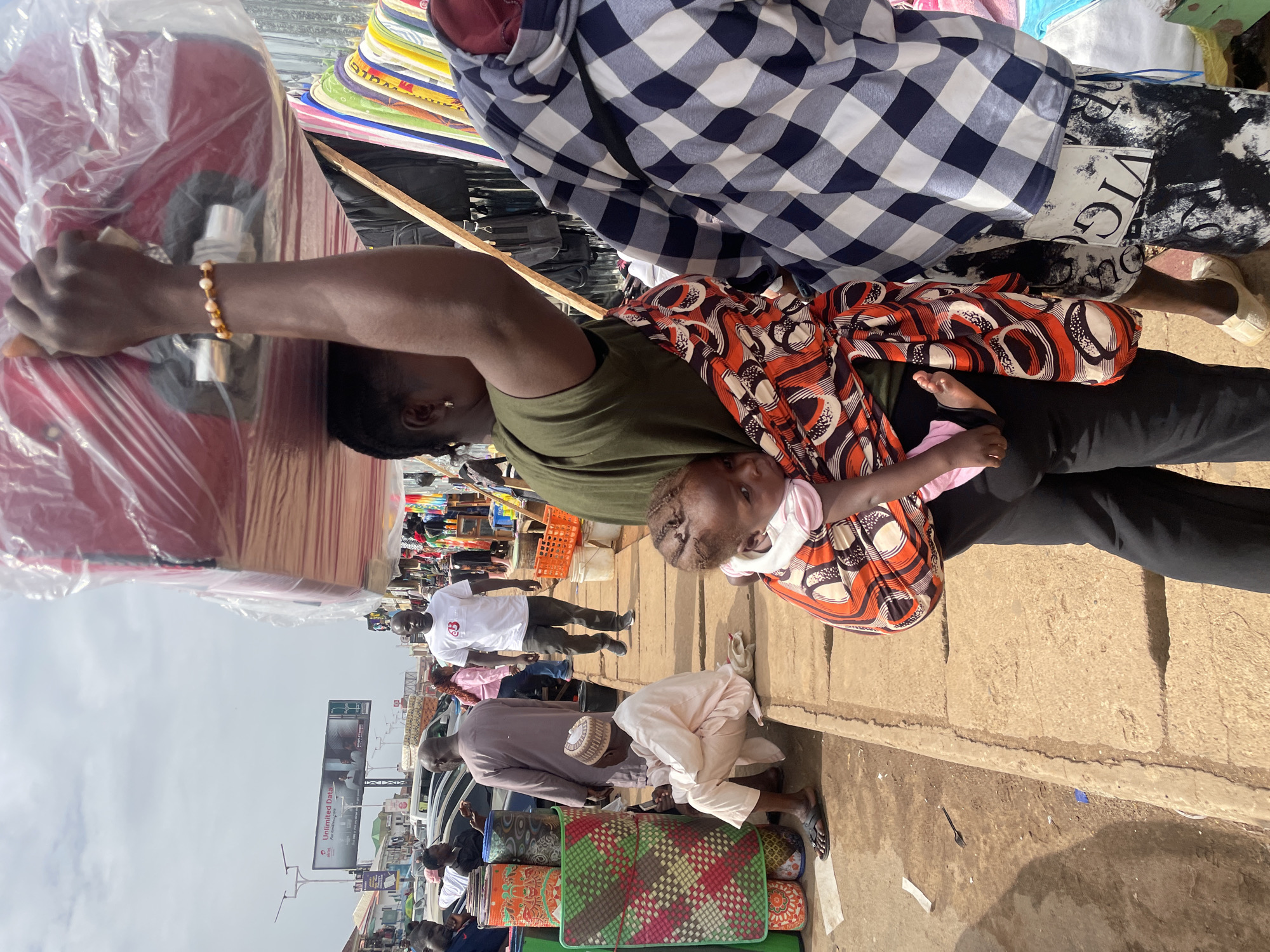 In Jos, Nigeria, an African mother carries a baby in a patterned orange and white sling. She is holding a stack of red and white items wrapped in plastic on her head, possibly for sale. The woman is wearing a green shirt and black pants. Several people walk along a busy street. A man bends down near mats, and billboards are visible in the background.