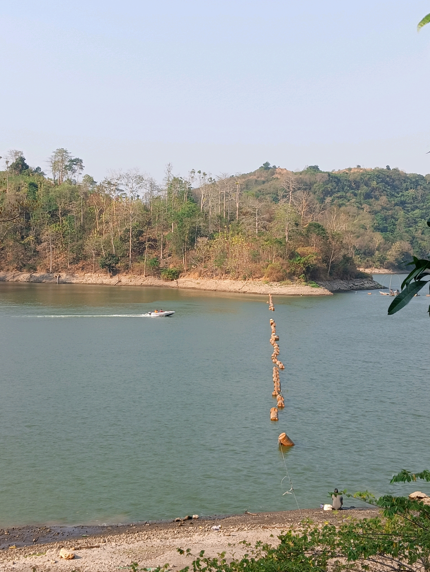 A wide shot captures a serene natural landscape, likely near Kandangmas, Indonesia, dominated by a vast body of calm, blue-green water. A white speedboat with two occupants is speeding across the water on the left side of the frame, leaving a white wake behind it. A line of orange buoys stretches diagonally across the water from the foreground towards the background, marking a boundary or path. The shoreline on the right is rocky and sparsely vegetated, with a single person sitting on the rocks, facing the water. In the background, a densely forested hillside rises, covered in a mix of green and bare trees, with a clear, pale sky above. The overall impression is one of tranquility and natural beauty.