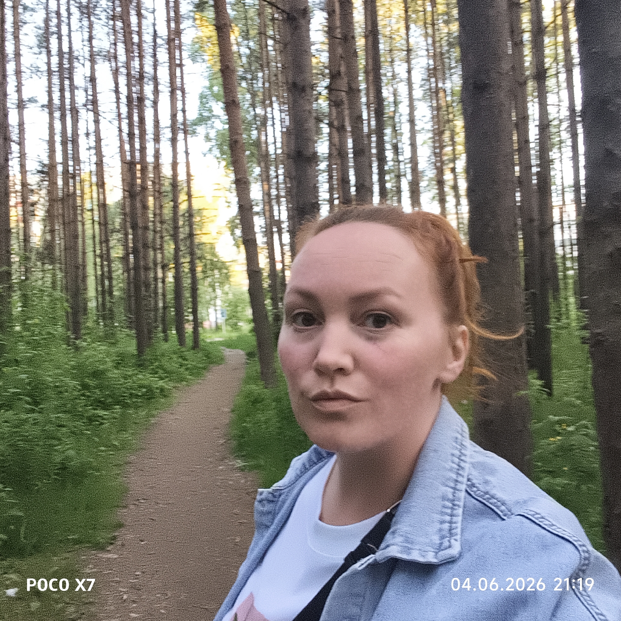 A woman is depicted in a selfie, positioned in the foreground of a forest scene in Chaikovsky, Russia. She has reddish-brown hair, is wearing a light blue denim jacket over a white t-shirt, and looks directly at the camera with a neutral expression.
The background is dominated by numerous tall, slender trees, likely pines or spruces, creating a dense woodland environment. A dirt path winds into the distance on the left side of the frame, flanked by lush green undergrowth. The lighting indicates late evening twilight, with warm, golden light filtering through the trees, consistent with the time stamp.
Visible text includes "POCO X7" in the bottom left corner, indicating the camera model, and "04.06.2026 21:19" in the bottom right, showing the date and time the photograph was taken. The scene evokes a tranquil, natural setting at dusk.