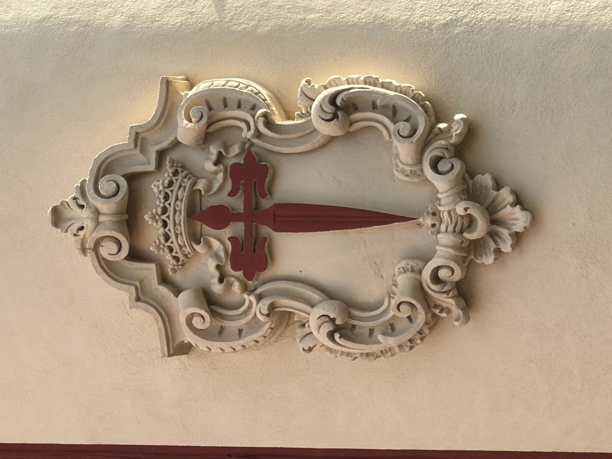 This image shows a close-up, eye-level view of a decorative architectural element featuring a crest. The crest is ornate and sculpted, with a light cream-colored finish, set against a textured, pale yellow wall. The crest is framed by elaborate scrollwork and topped with a crown. In the center of the crest is a prominent red cross with a sword-like shape embedded within it. The lighting suggests it is daytime, with sunlight casting shadows that highlight the three-dimensional details of the sculpture. A sliver of a dark red vertical line is visible on the far left edge of the frame, possibly part of a doorway or window frame. The overall impression is of historical ornamentation found on a building. The crest is that of the Order of Santiago, and this particular emblem is located on the facade of the Palacio de Congresos de Palma, also known as the Palma Auditorium, in Palma de Mallorca, Spain.