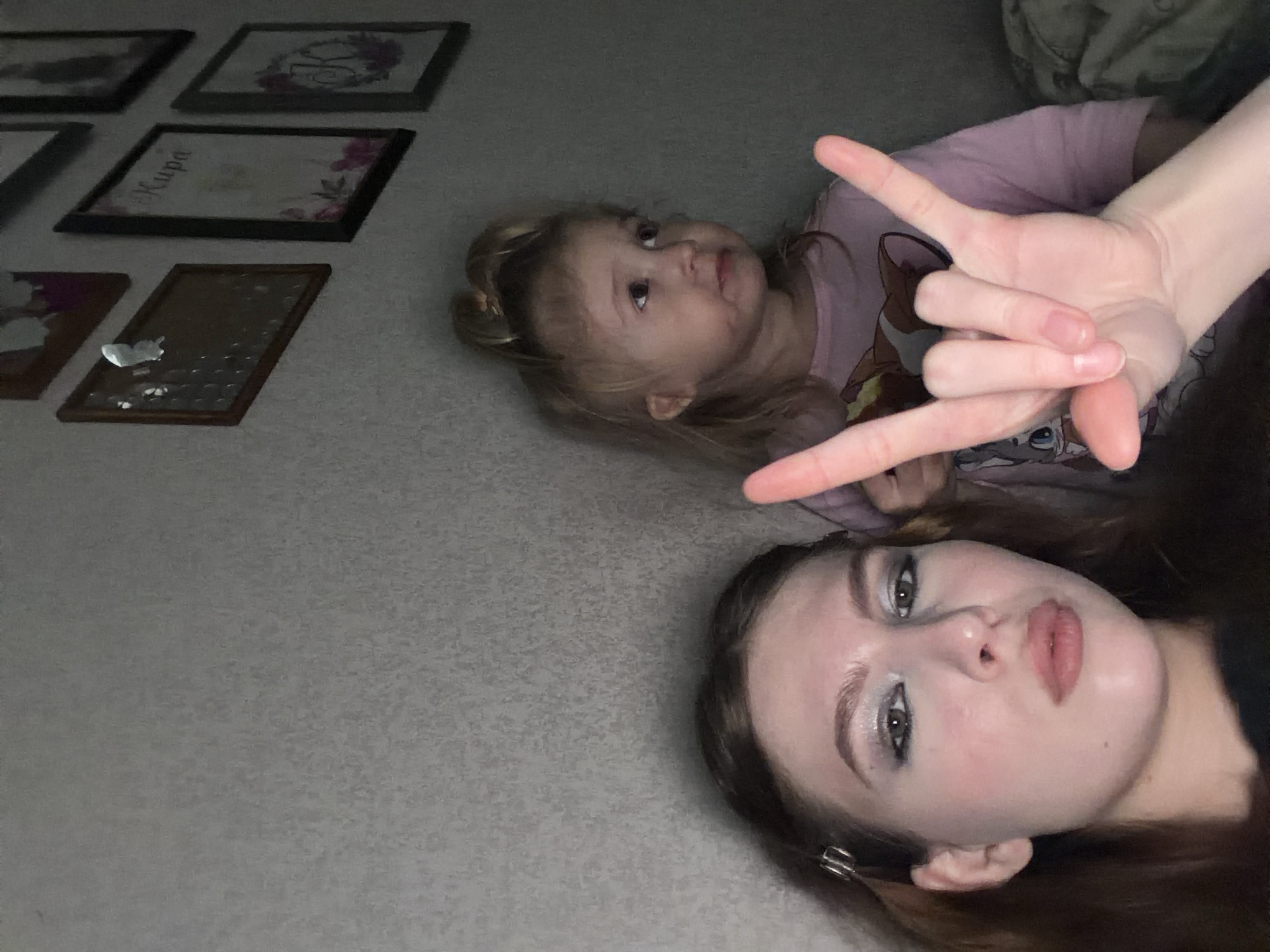 Two individuals, one appearing to be a young girl and the other a teenage girl or young woman, are lying on what appears to be a carpeted floor. The younger girl is wearing a pink t-shirt with a cartoon design. The older girl/young woman is wearing dark clothing and has makeup applied. A hand is positioned in the foreground making a hand gesture. Several framed pictures are visible on the wall in the background. The location is Noviy, Russia.