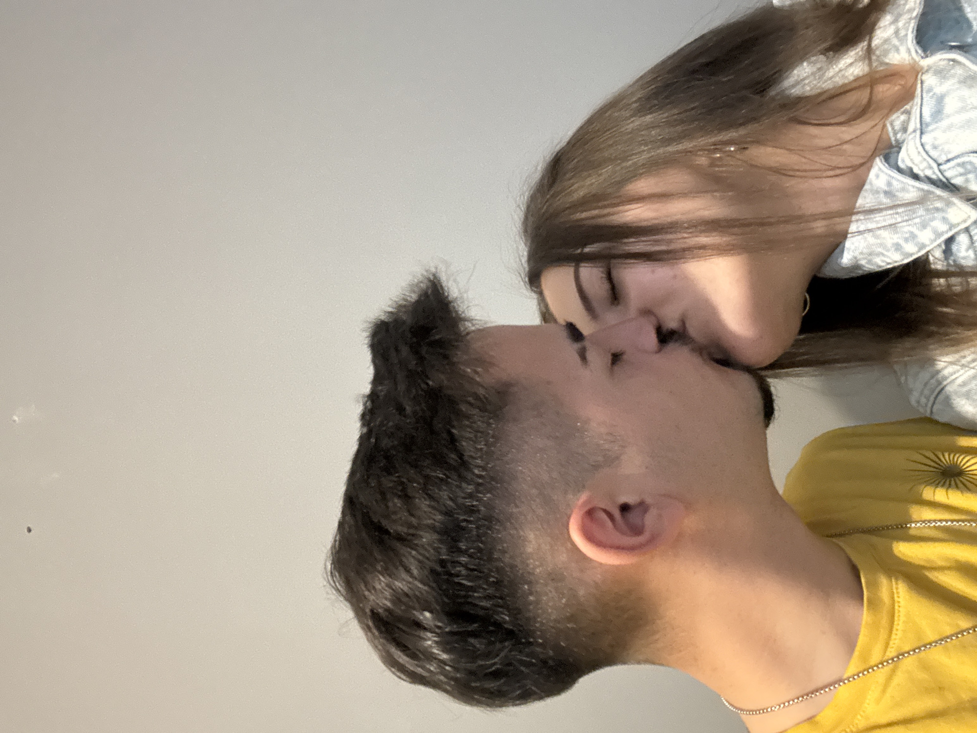 Two people are in the image, appearing to be in an embrace and sharing a kiss. One person is wearing a yellow t-shirt with a sun-like design and a thin gold chain around their neck, and their dark hair is cut short on the sides. The other person is wearing a denim jacket. The background is a plain, light-colored wall. The image does not contain information that can identify the location in the city of Sofia, Bulgaria.
