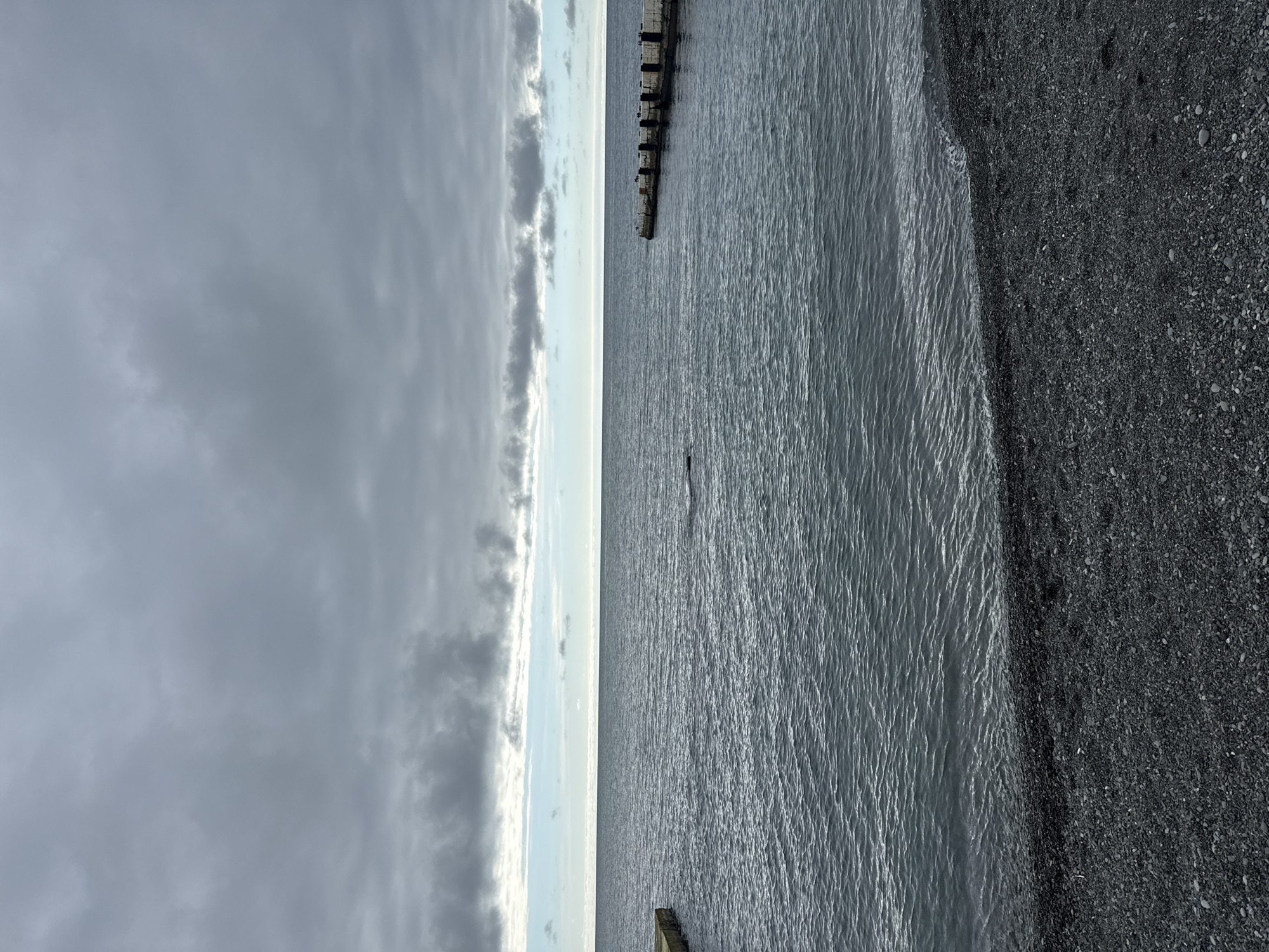 This image depicts a coastal scene in Sochi, Russia, under an overcast sky. The upper half of the frame is dominated by a dense, grey cloud cover, with a brighter, light blue-grey band visible along the distant horizon line, where the sky meets the sea. The sea itself is a deep blue-grey, exhibiting a textured, rippled surface, indicative of slight wave activity. In the foreground, a dark grey pebble beach curves gently along the shoreline, meeting the water with a soft edge where small waves are breaking.
Two concrete breakwater structures extend into the water. One is partially visible on the far left of the frame, appearing as a truncated concrete wall. The second, more prominent structure is located on the right side, extending from the unseen shoreline into the sea. This breakwater is composed of a series of dark, rectangular concrete blocks forming a linear barrier, with several small, dark avian figures perched intermittently along its upper surface. No individuals or vessels are discernible within the frame.