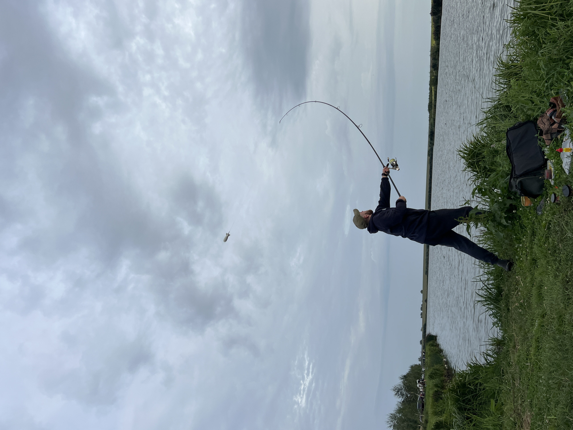 A man is fishing on the bank of a lake in Laskivtsi, Ukraine. He is wearing a dark hooded jacket, dark pants, and a green baseball cap. He is holding a fishing rod with the line cast into the water. The sky is overcast with clouds. In the distance, there are trees and some parked cars. A fishing bag and other fishing gear are on the ground next to the man.