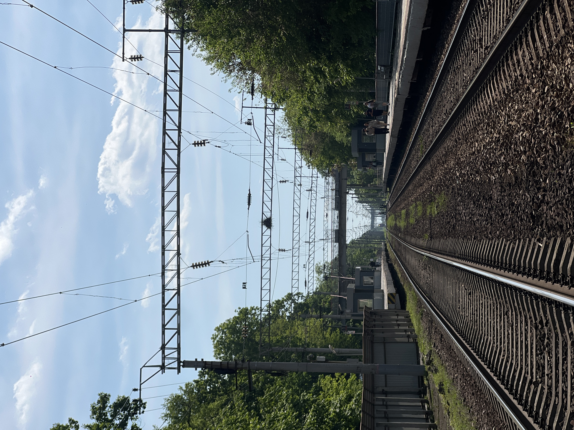 The image depicts a railway scene with tracks extending into the distance. Overhead power lines and support structures are visible above the tracks, interspersed with trees. A platform is located to one side of the tracks. People stand on the platform. The scene is set under a blue sky with scattered clouds. The location is likely Vladivostok, Russia.