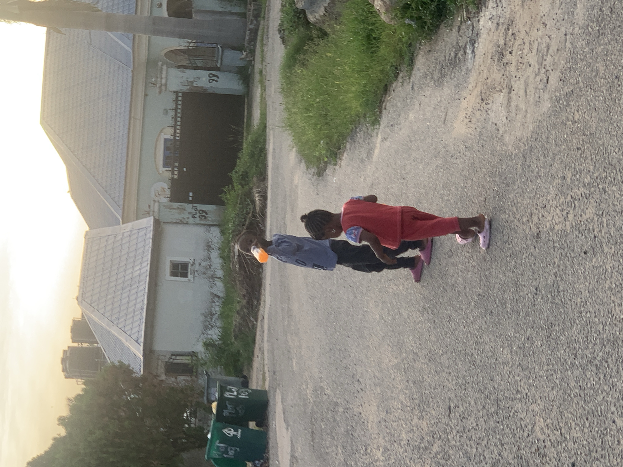 Two children are walking on a paved road. One child is wearing a red top and pants with pink sandals; the other is wearing a blue shirt and black pants with pink sandals, holding an orange plastic bottle. Green grass and vegetation line the side of the road. In the background are buildings, including one with the number "99" on it, and green recycling bins. The photo was taken at dusk or dawn. The location is likely a residential area in Abuja, Nigeria, based on architectural style, vegetation, and recycling bin markings.