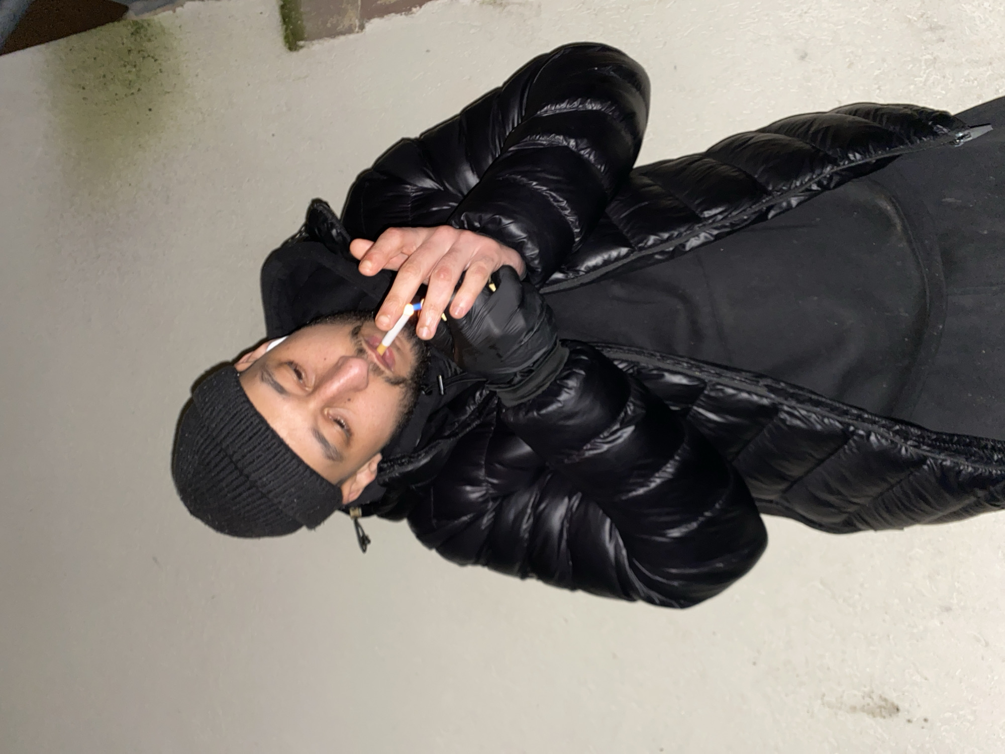 A man wearing a black beanie, a black puffer jacket, and gloves is smoking a cigarette. He is situated on a concrete surface, possibly a sidewalk. The location may be Schiltigheim, France.